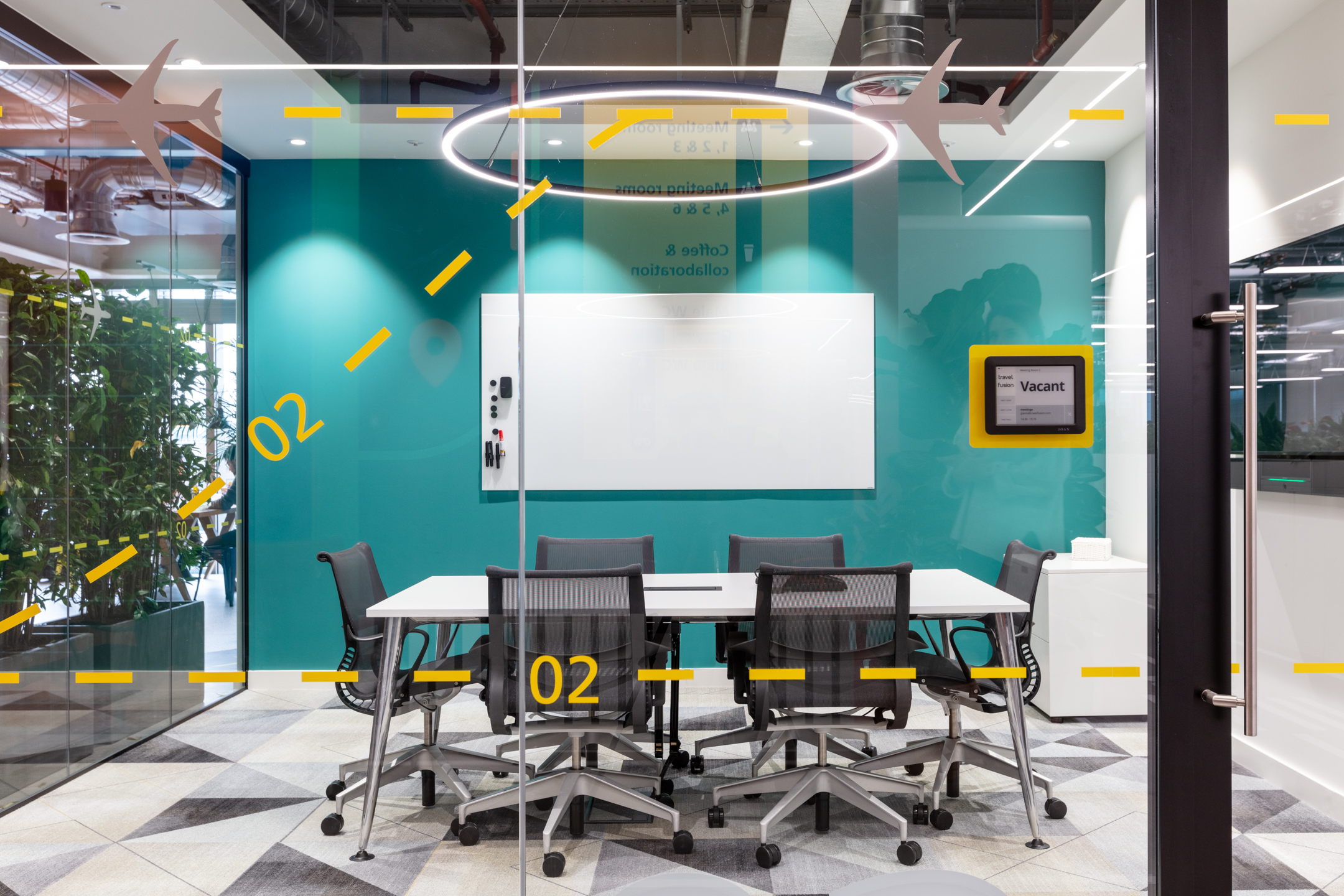 Travelfusion’s modern meeting room with glass walls, featuring a central table, office chairs, and a teal accent wall with a whiteboard. The glass walls have yellow aviation-themed markings, and a digital sign indicates the room is