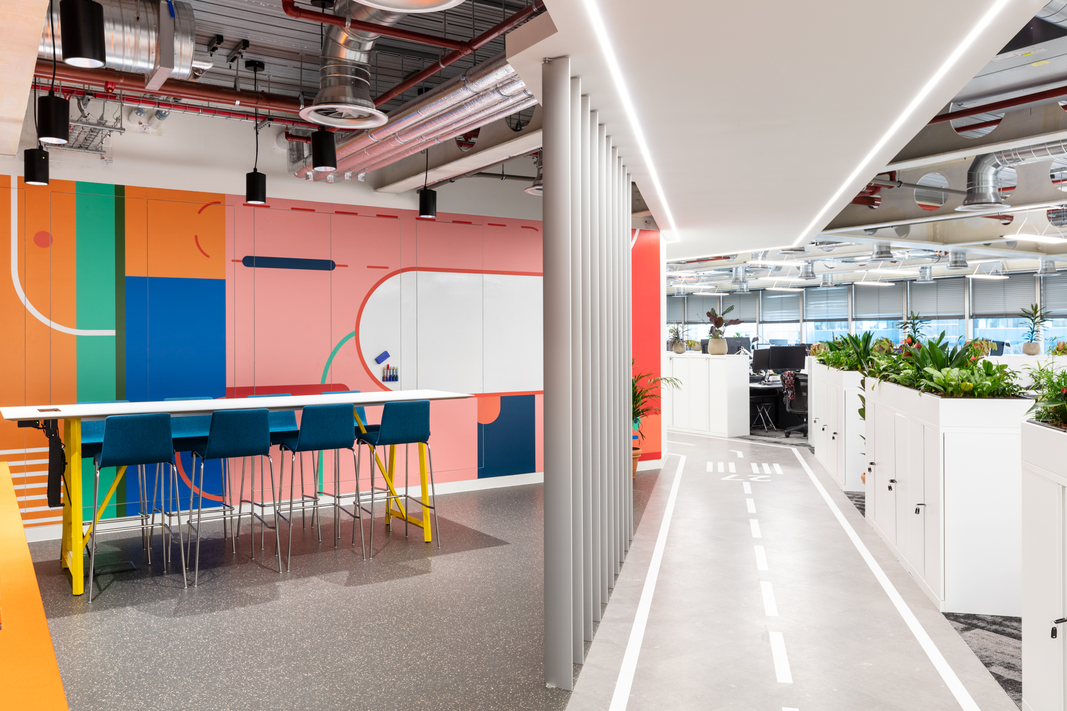 Travelfusion’s vibrant workspace featuring a colourful, geometric wall with bold patterns, a high-top table, and exposed ductwork overhead. A pathway resembling a road runs alongside the workstations, lined with plants and white storage units.