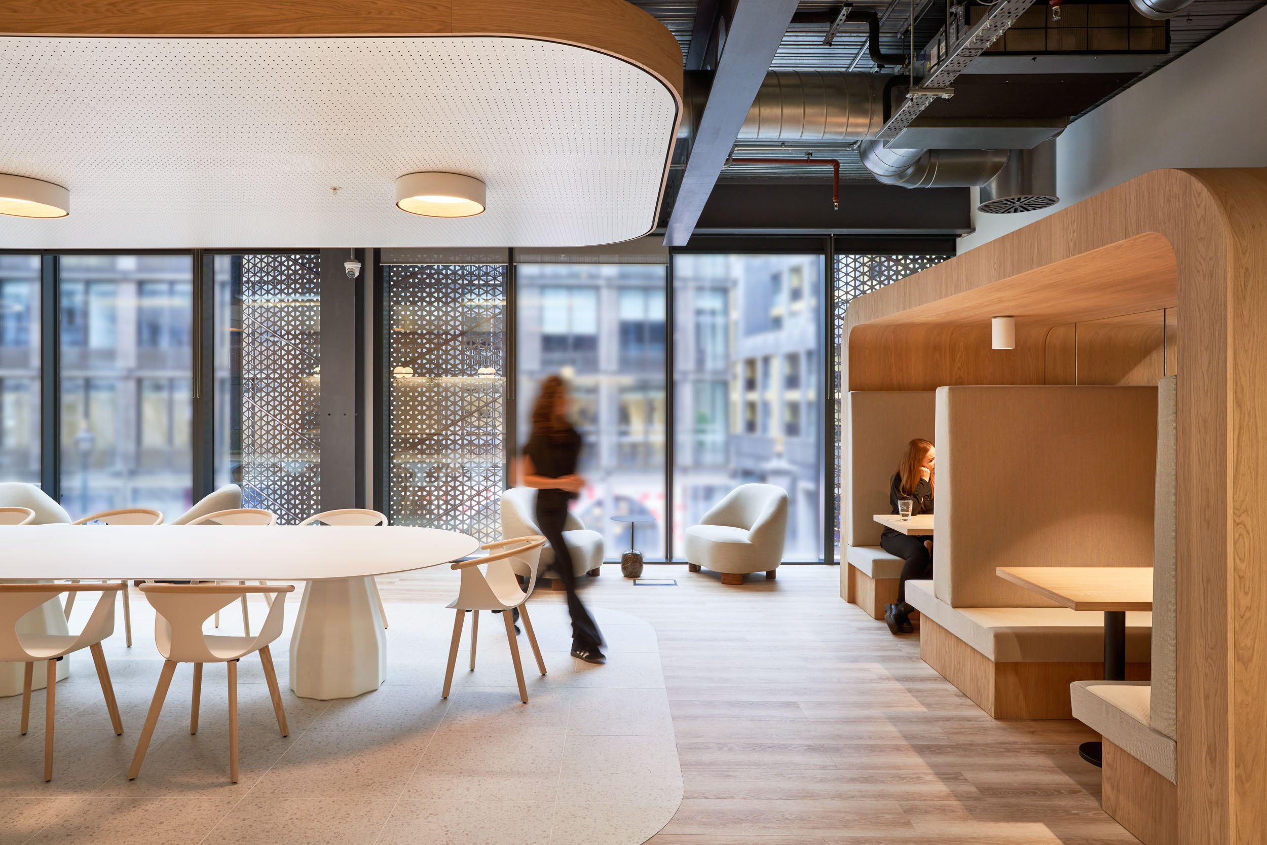 : Financial Services company office designed by Modus Workspace, featuring a modern collaboration space with a large oval table, comfortable seating, and built-in booths. A person is walking through the bright, naturally lit space, adding a sense of movement to the inviting atmosphere for meetings and focused work