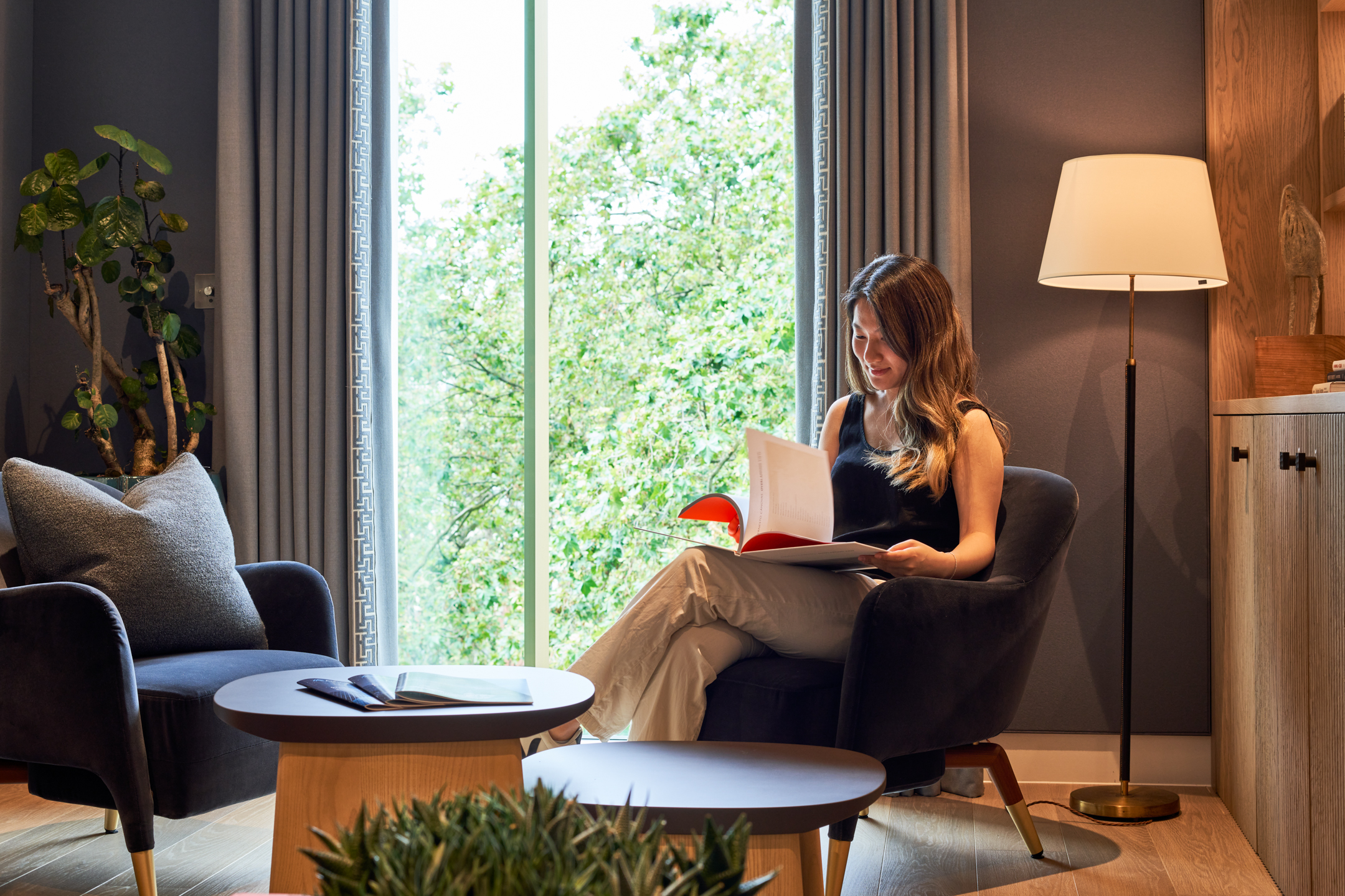 Asset Management Firm office designed by Modus Workspace, featuring a calm, relaxing seating area with a woman reading in a comfortable chair near a large window. The space is warmly lit with natural light, accented by greenery, soft furnishings, and a standing lamp, creating a serene environment for focused work.