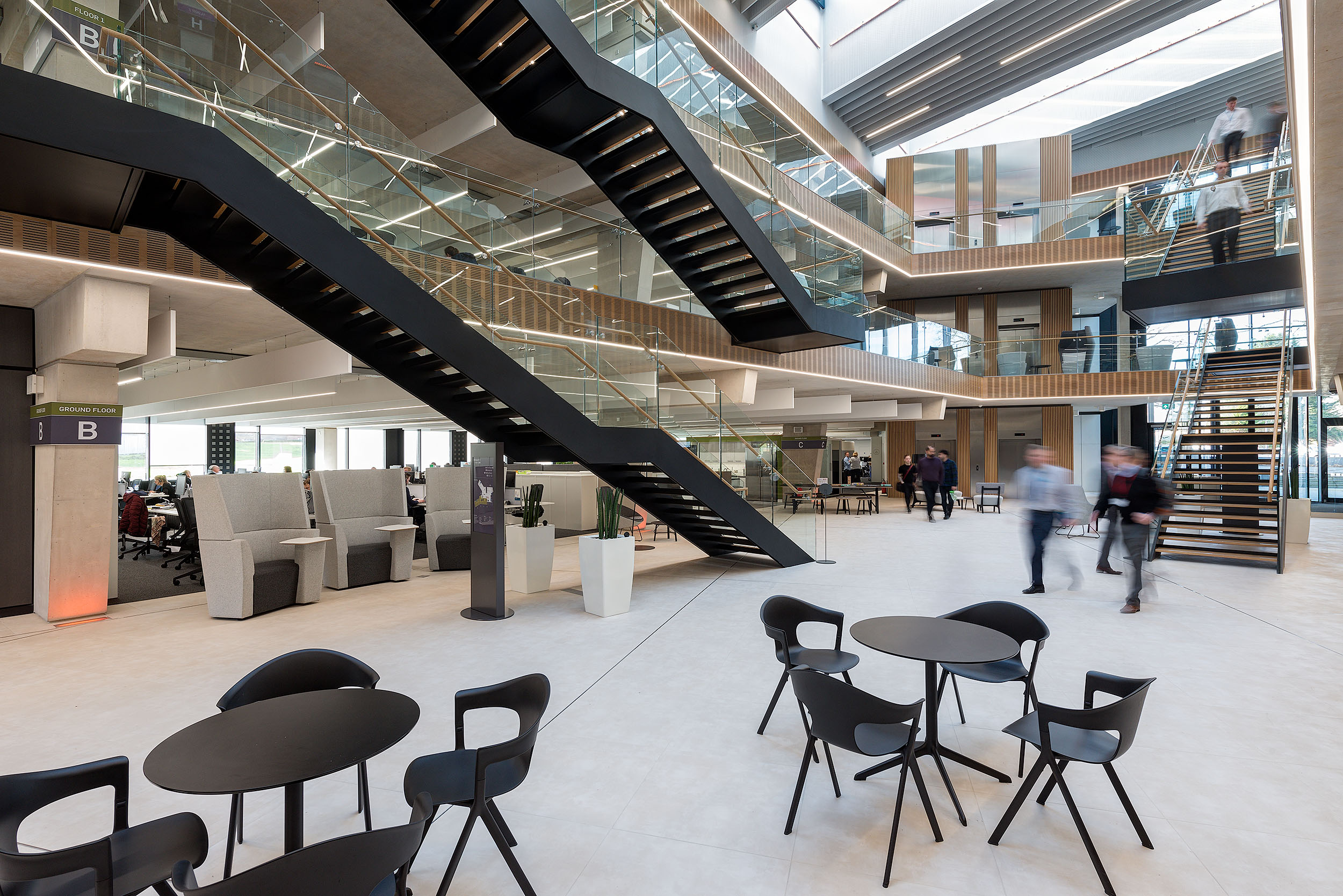 A view of the main atrium at Atkins Woodcote Grove with black staircases connecting the levels, surrounded by seating areas and open workspace zones.