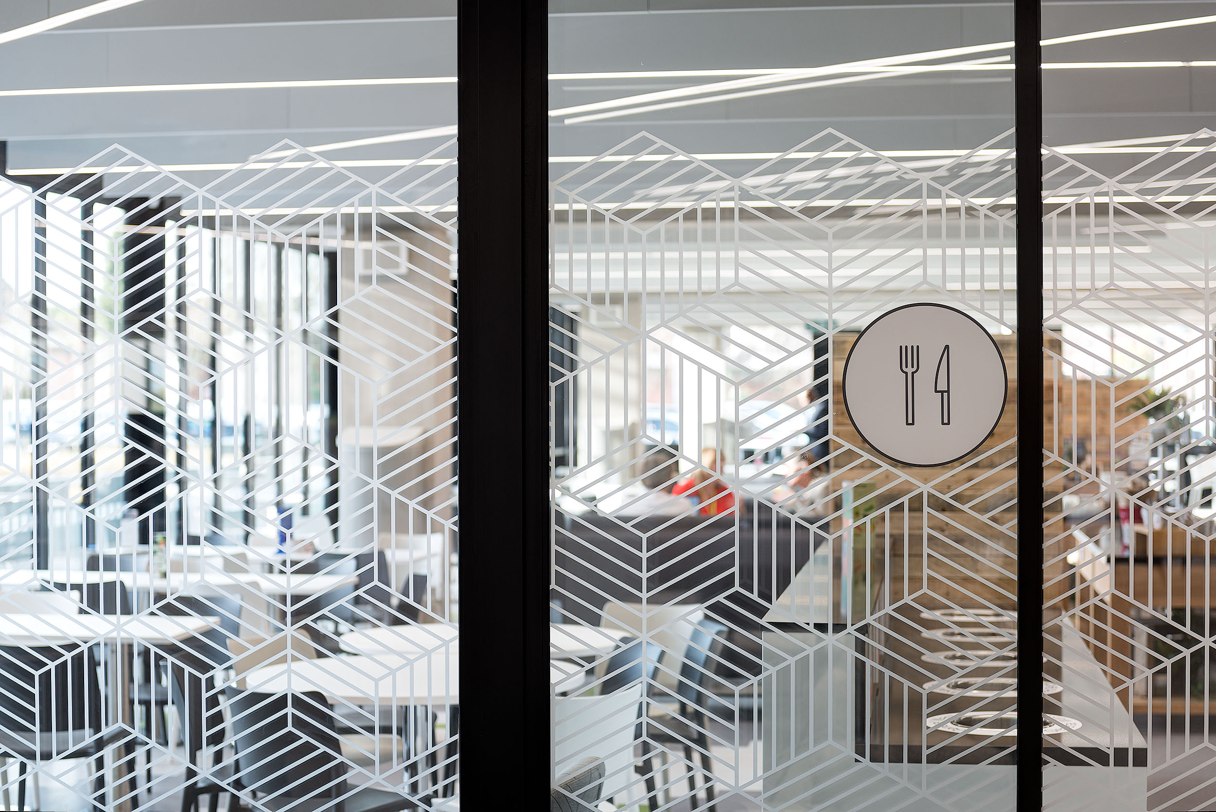 A close-up of a glass door with geometric patterns leading into a cafeteria at Atkins Woodcote Grove, with minimalistic design.