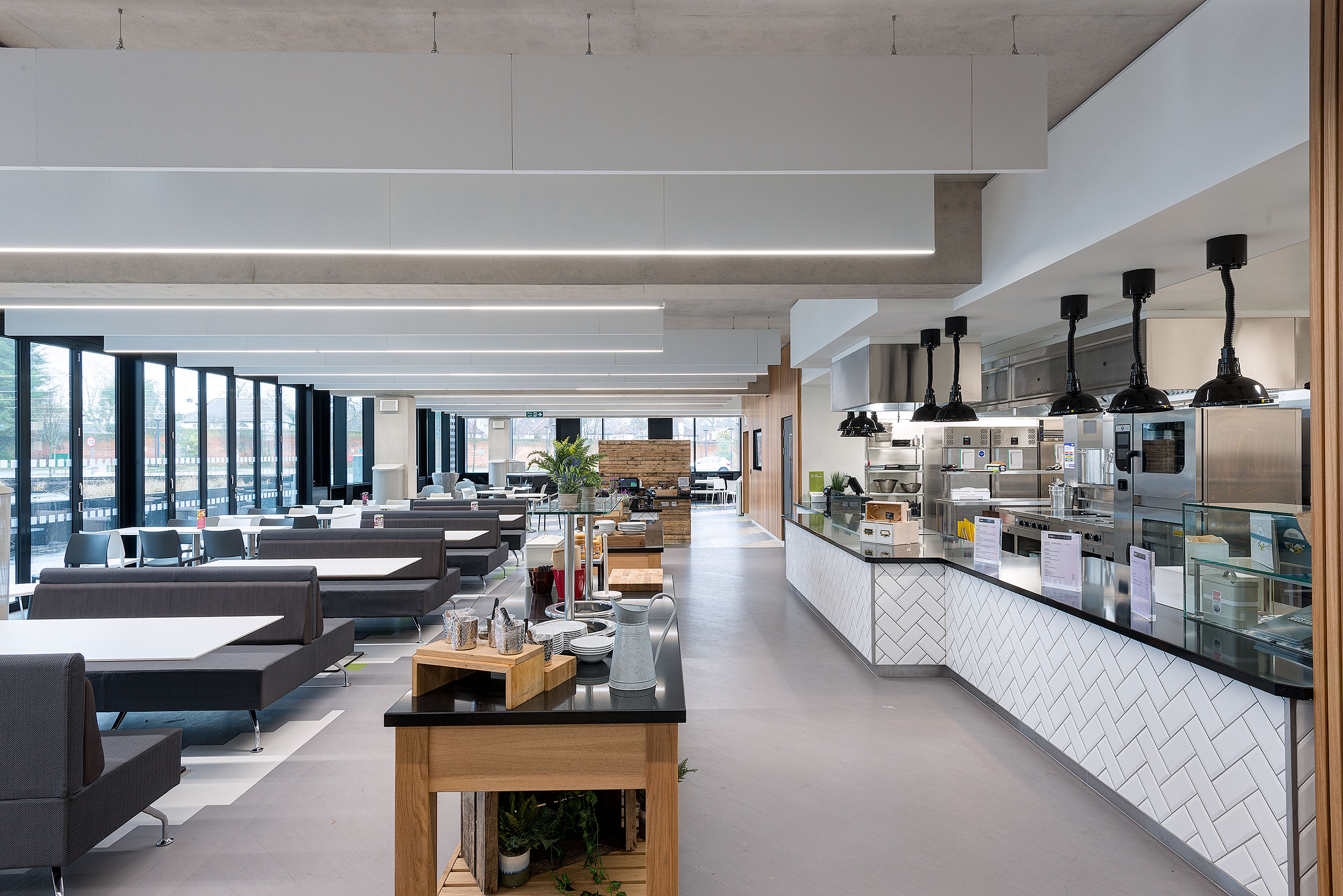 A spacious cafeteria at Atkins Woodcote Grove, with large windows, booth seating, and a bright, open kitchen area.