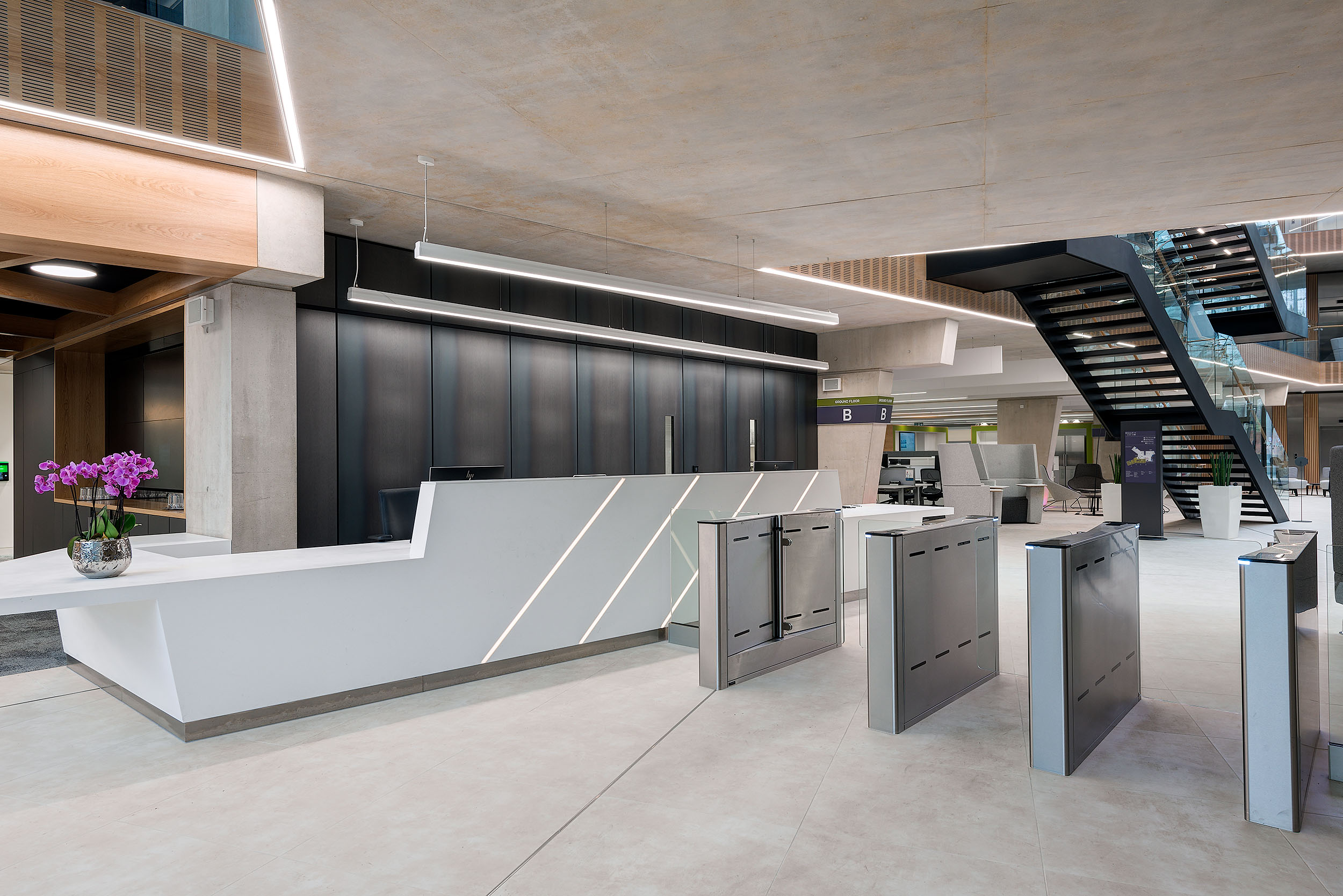The reception area at Atkins Woodcote Grove features a modern white desk, security barriers, and a staircase leading up from the lobby.