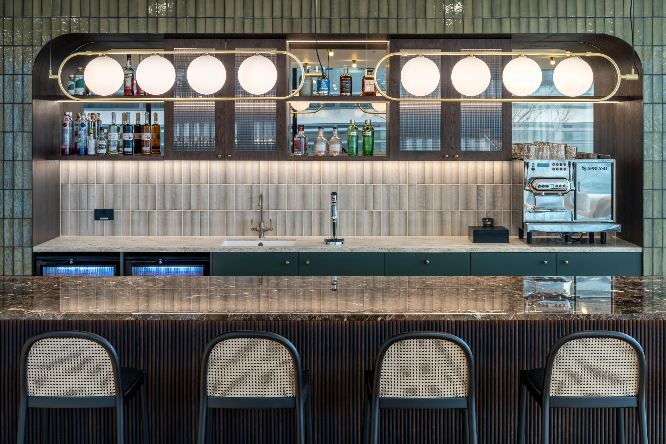 A close-up of the bar at Authentic Brand Group, designed by Modus, highlighting the elegant marble counter, textured tile backdrop, and stylish shelving with liquor bottles and a coffee machine.