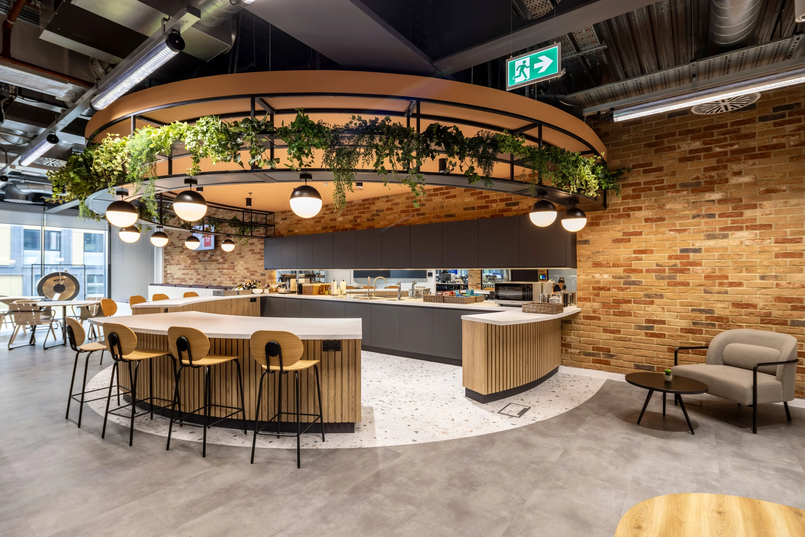 Bazaarvoice's London office kitchen area, designed by Modus Workspace, featuring modern seating and a hanging greenery installation. The exposed brick walls and warm lighting create a welcoming space for collaboration and relaxation.
