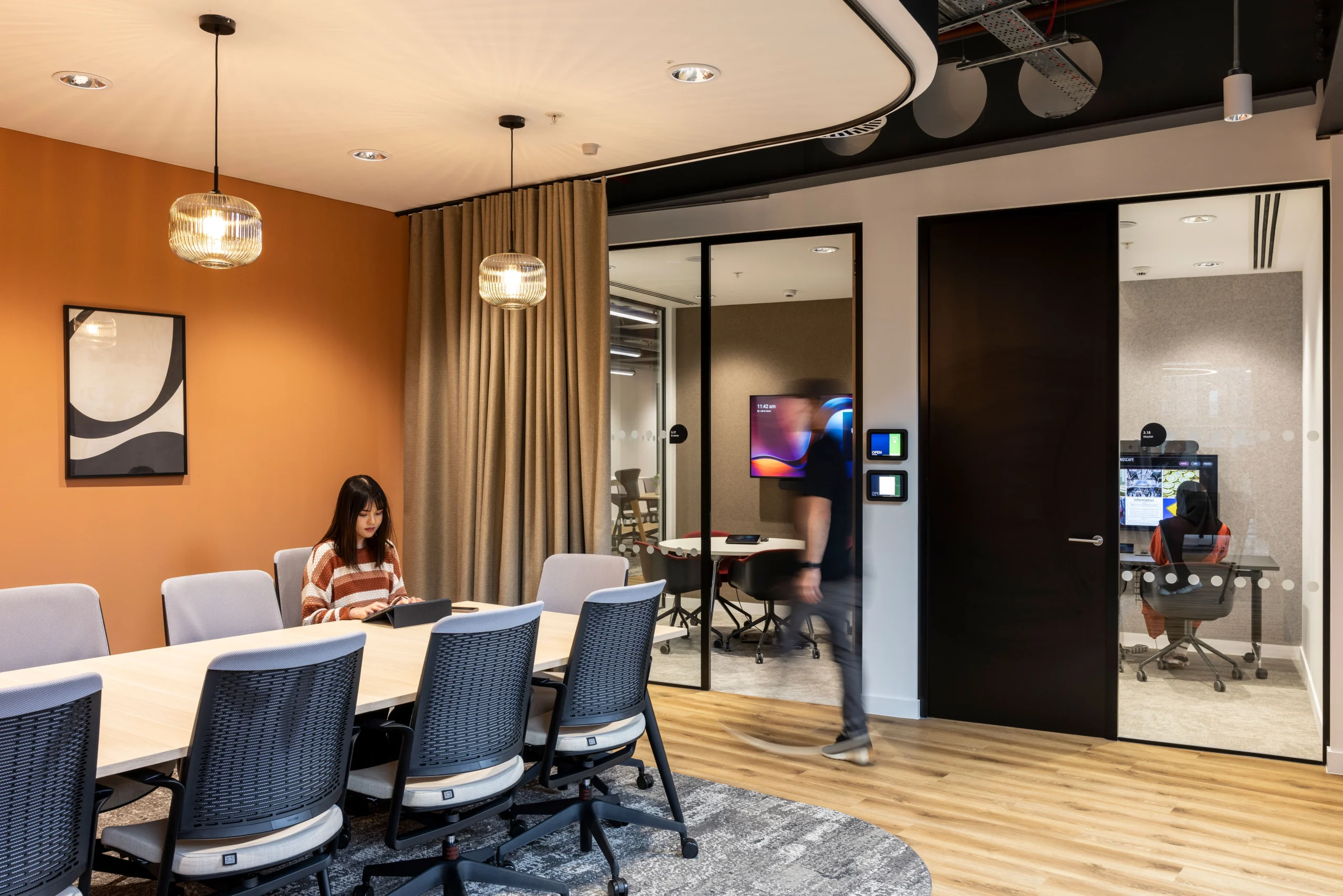 A modern meeting room at Bazaarvoice's London office, designed by Modus Workspace, where a team member is seen working diligently. The room features contemporary furniture, a striking orange accent wall, and advanced technology, creating an ideal space for strategic discussions and presentations.
