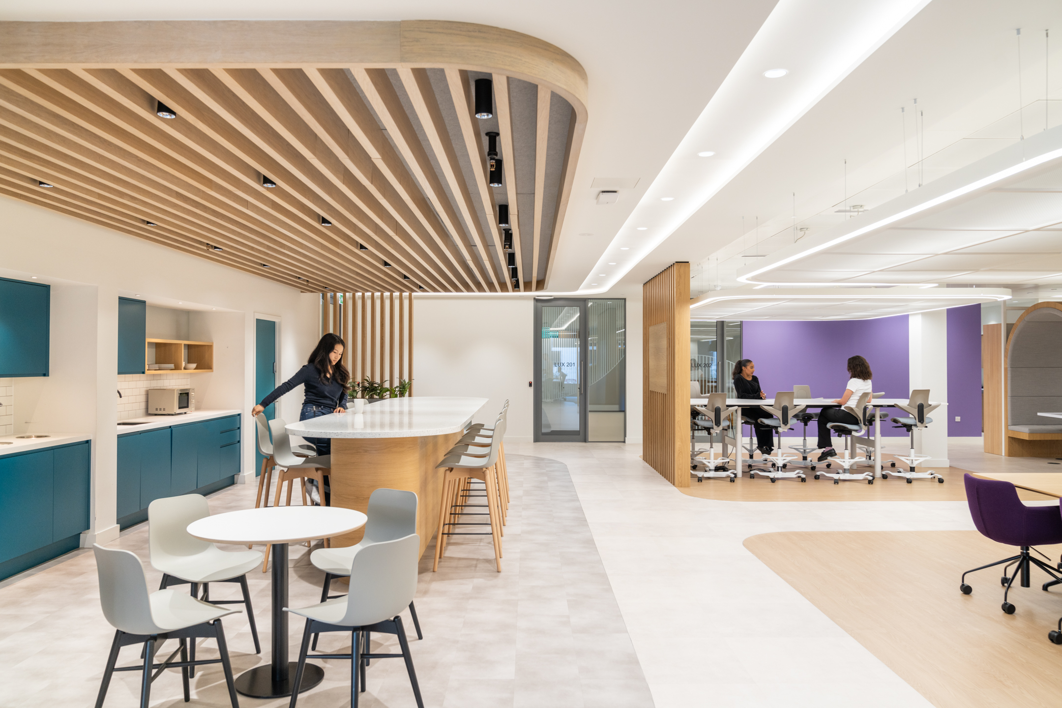 DXC Technology collaborative workspace and teapoint feature a long island, highchairs, and individual meeting areas with teal accents and warm wood finishes.