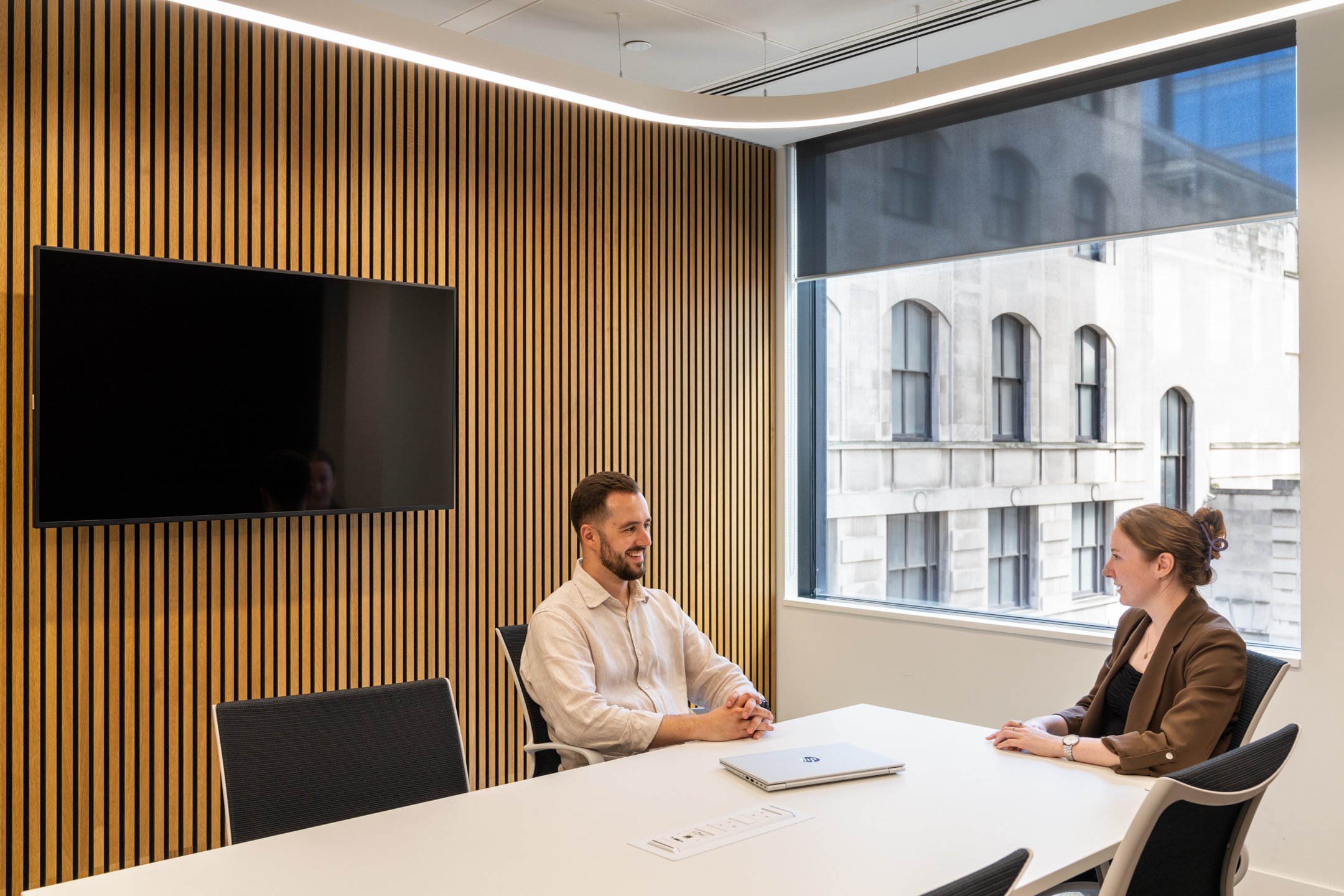 DXC Technology's small meeting room with wooden slatted walls, a large window, and two people seated at a white table facing a wall-mounted screen.