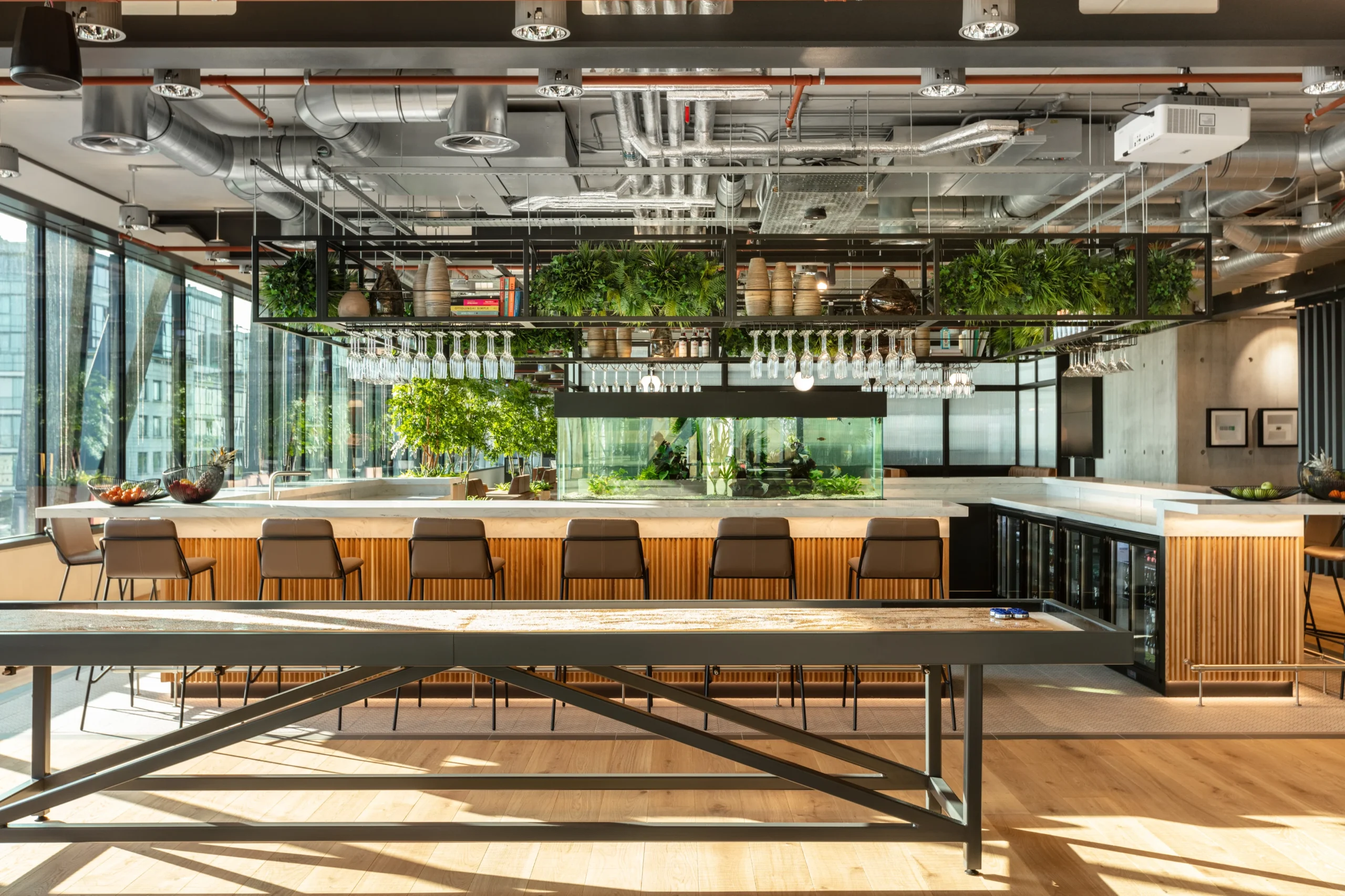 Bar area at Alpha FX, with a shuffleboard table in the foreground, a large counter with bar stools, overhead greenery, hanging glassware, and a built-in fish tank, illuminated by natural light from the surrounding windows.