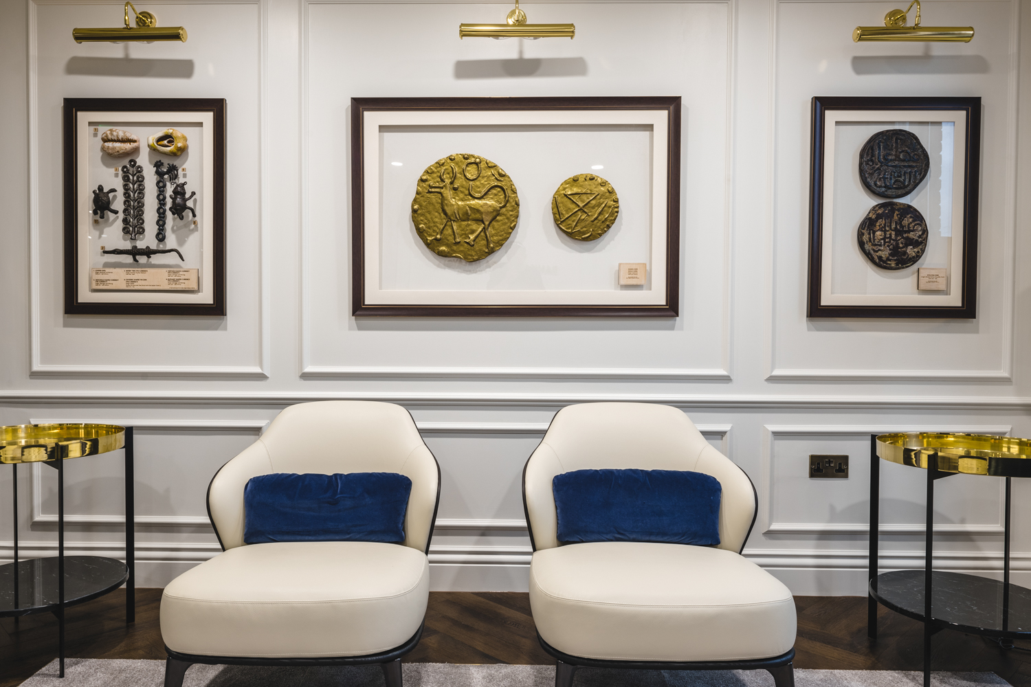 Two modern white lounge chairs with blue cushions, displayed against framed art pieces and gold lighting fixtures in the office.