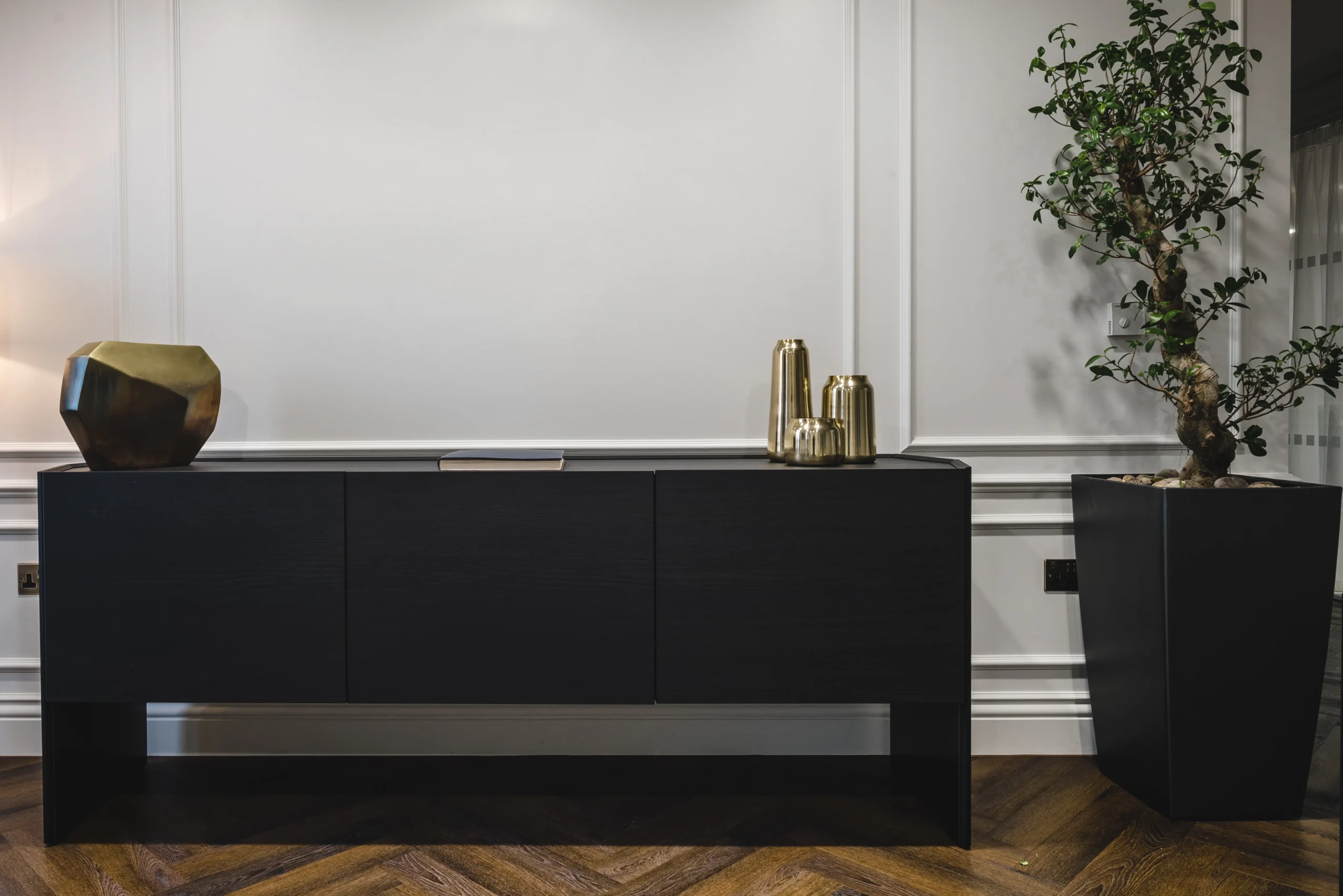 Black sideboard with gold decor and a potted plant on a wood herringbone floor, placed along the panelled walls of the bank's office.