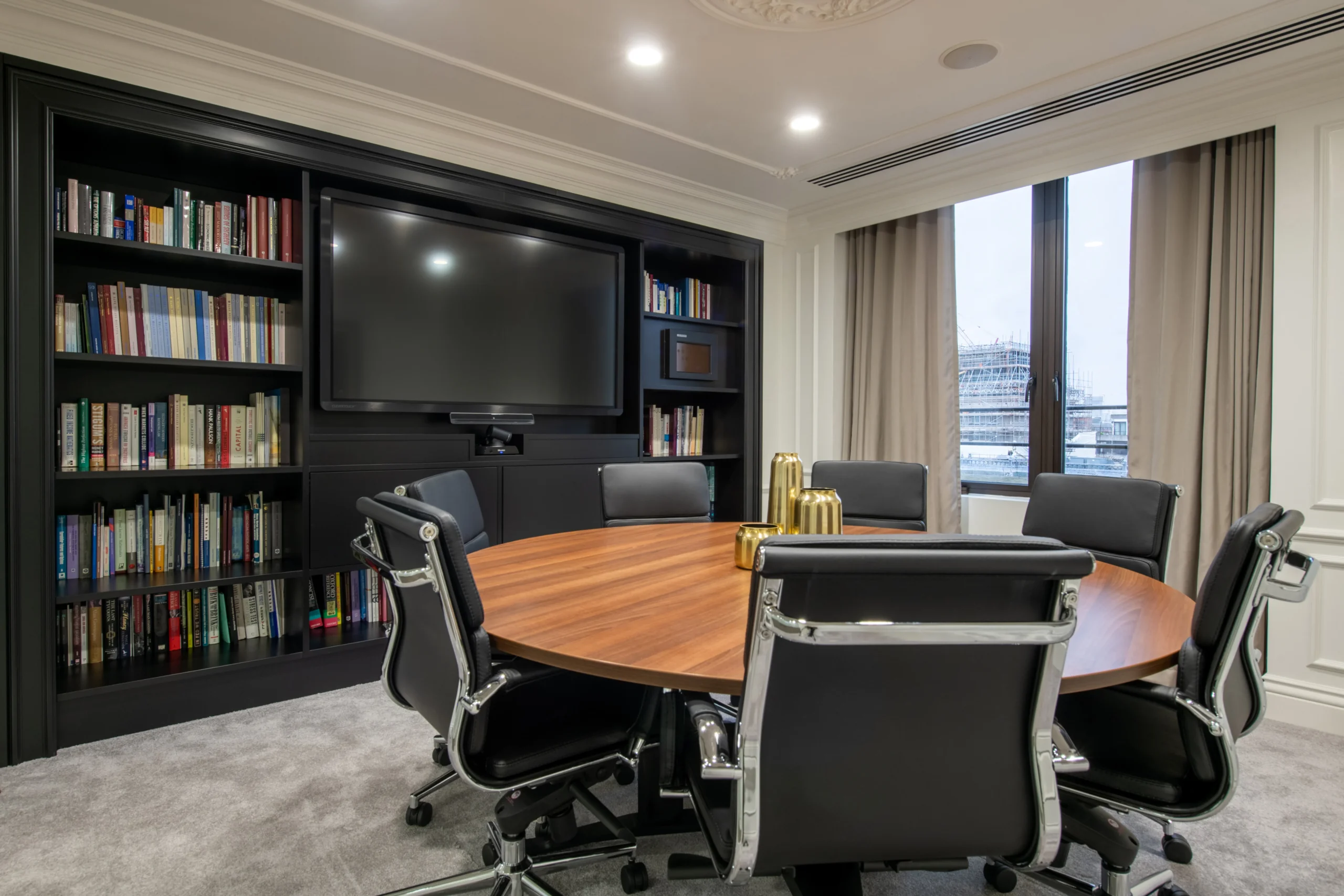 Small meeting room with a circular wooden table and black leather chairs, complemented by a built-in bookshelf and a large screen.