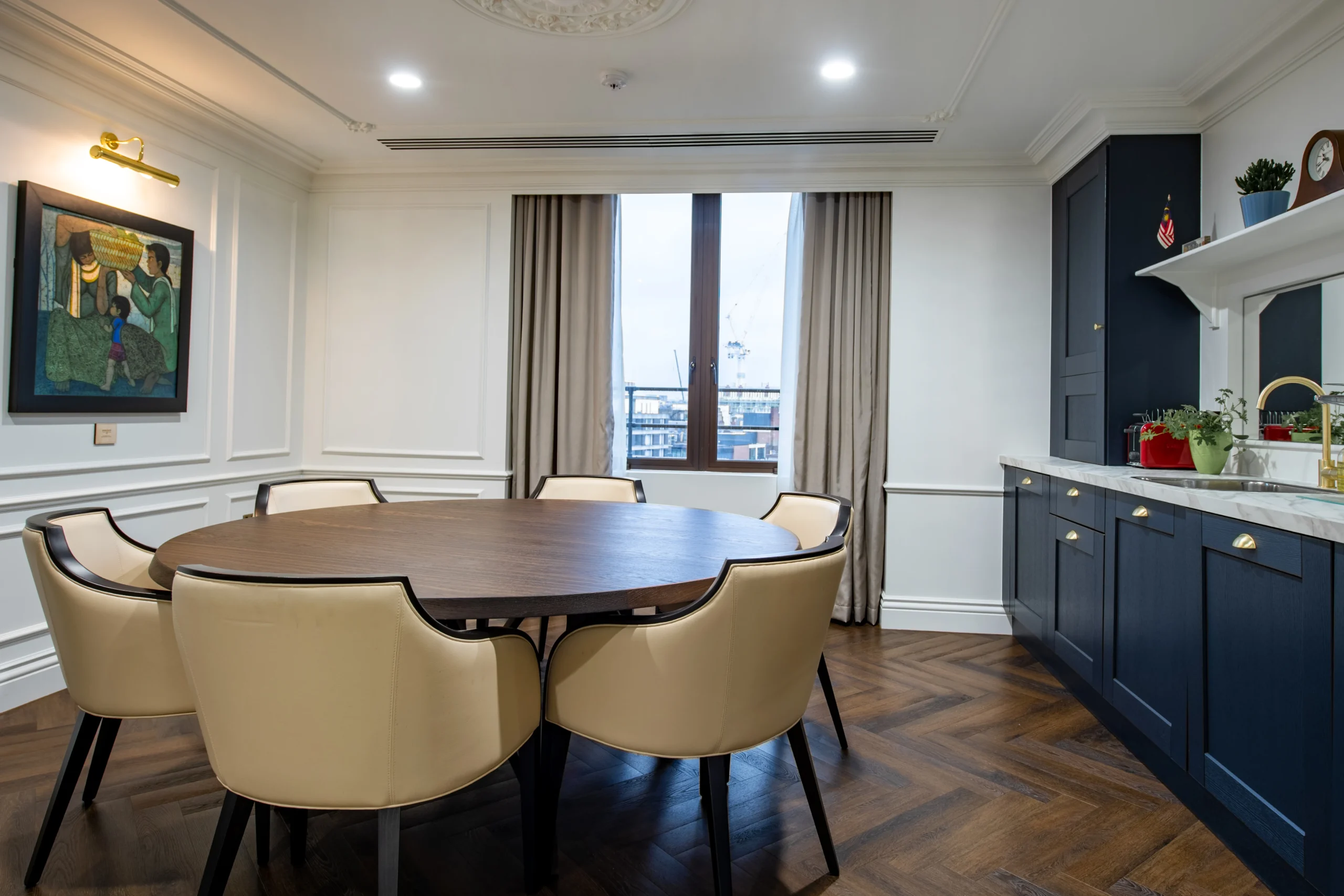 Small meeting area with beige leather chairs surrounding a round wooden table, placed near a built-in kitchenette with navy blue cabinets.