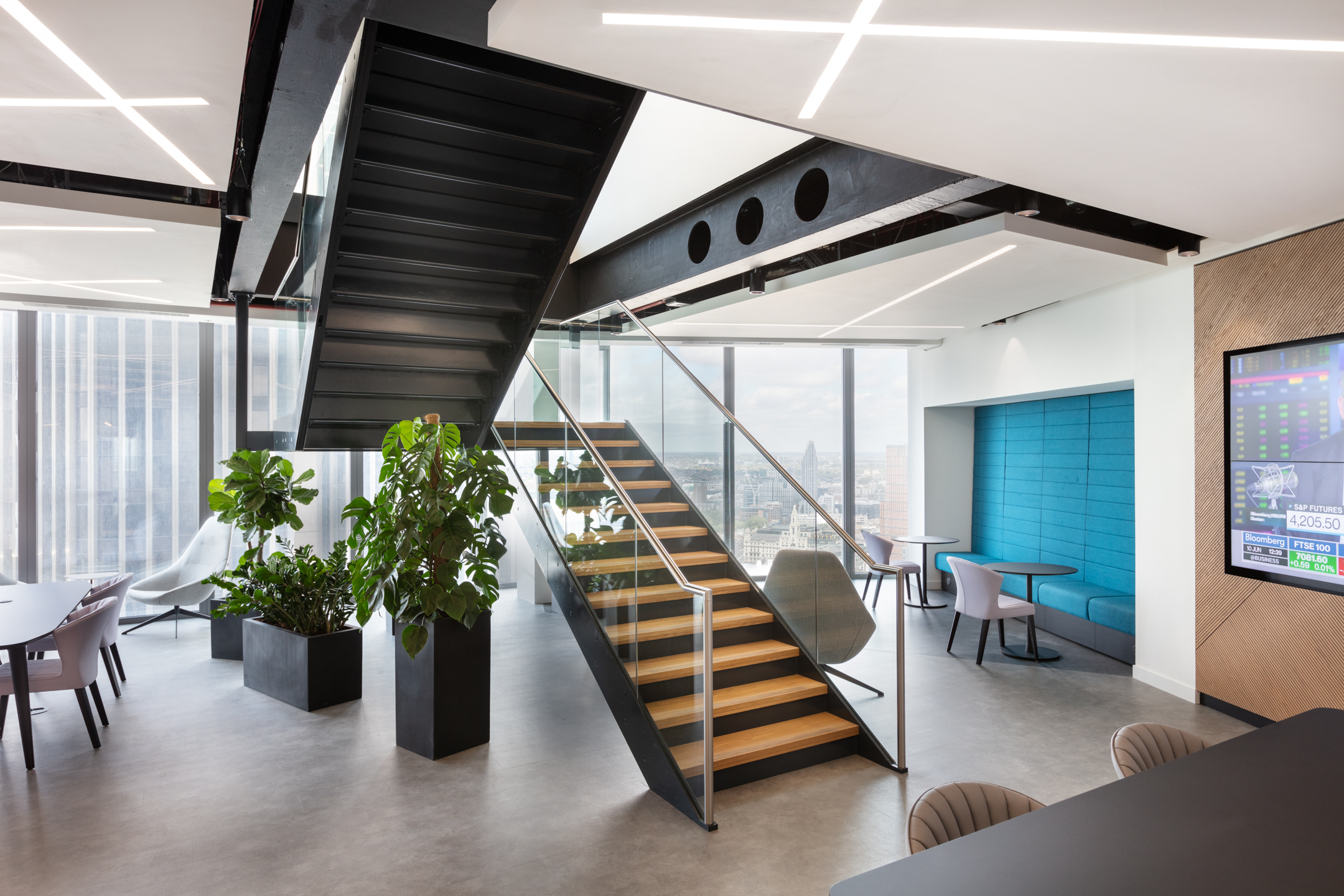 A black steel staircase with wooden steps and glass railings connects two floors of the office, surrounded by floor-to-ceiling windows and soft seating at the Insurance and Risk Management Firm in Bishopsgate.