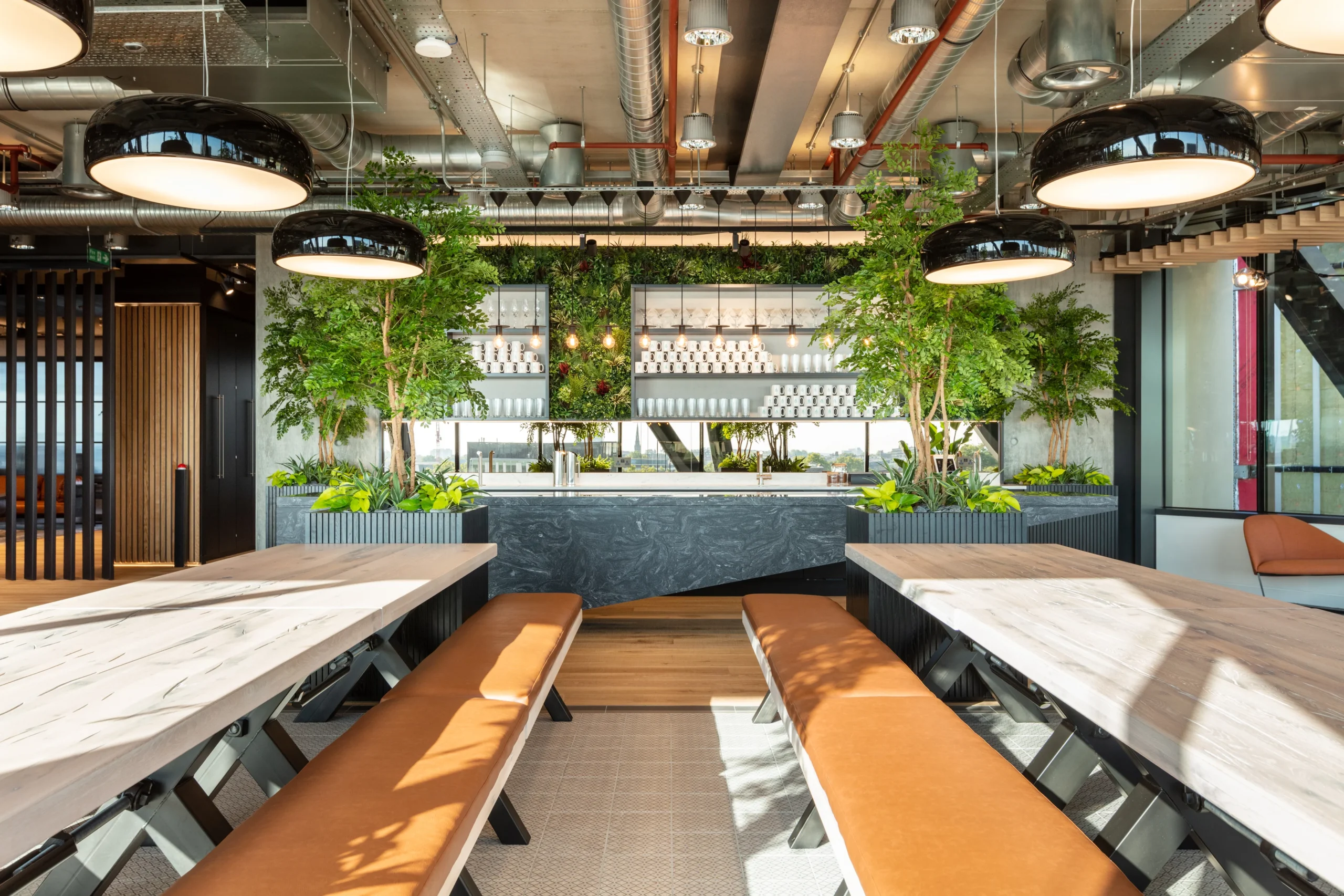 A modern teapoint and dining area in the Alpha FX office, designed by Modus Workspace, featuring long wooden tables with bench seating and leather cushions. The space is complemented by an industrial-style bar with a biophilic living wall integrated into the design, exposed ceilings, and large potted plants, creating a blend of natural elements and industrial aesthetics.