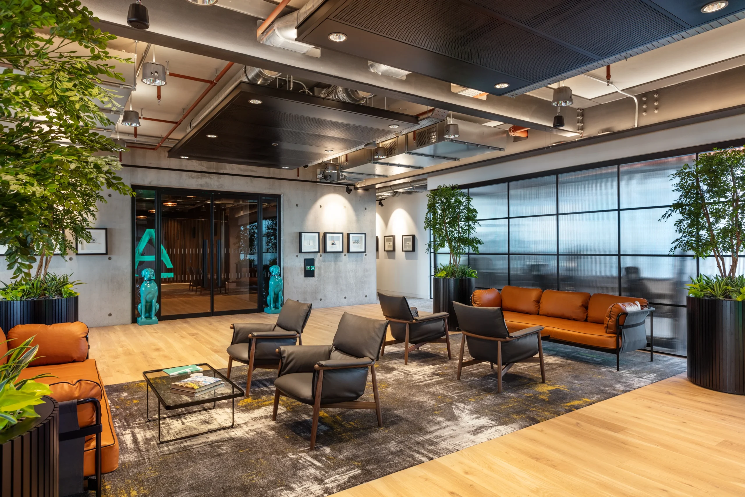 A stylish office lounge for Alpha FX, designed by Modus Workspace, featuring leather sofas and modern armchairs arranged on a textured rug. The space has an industrial aesthetic with exposed ceilings, concrete walls, and large potted plants, creating a sophisticated yet comfortable environment