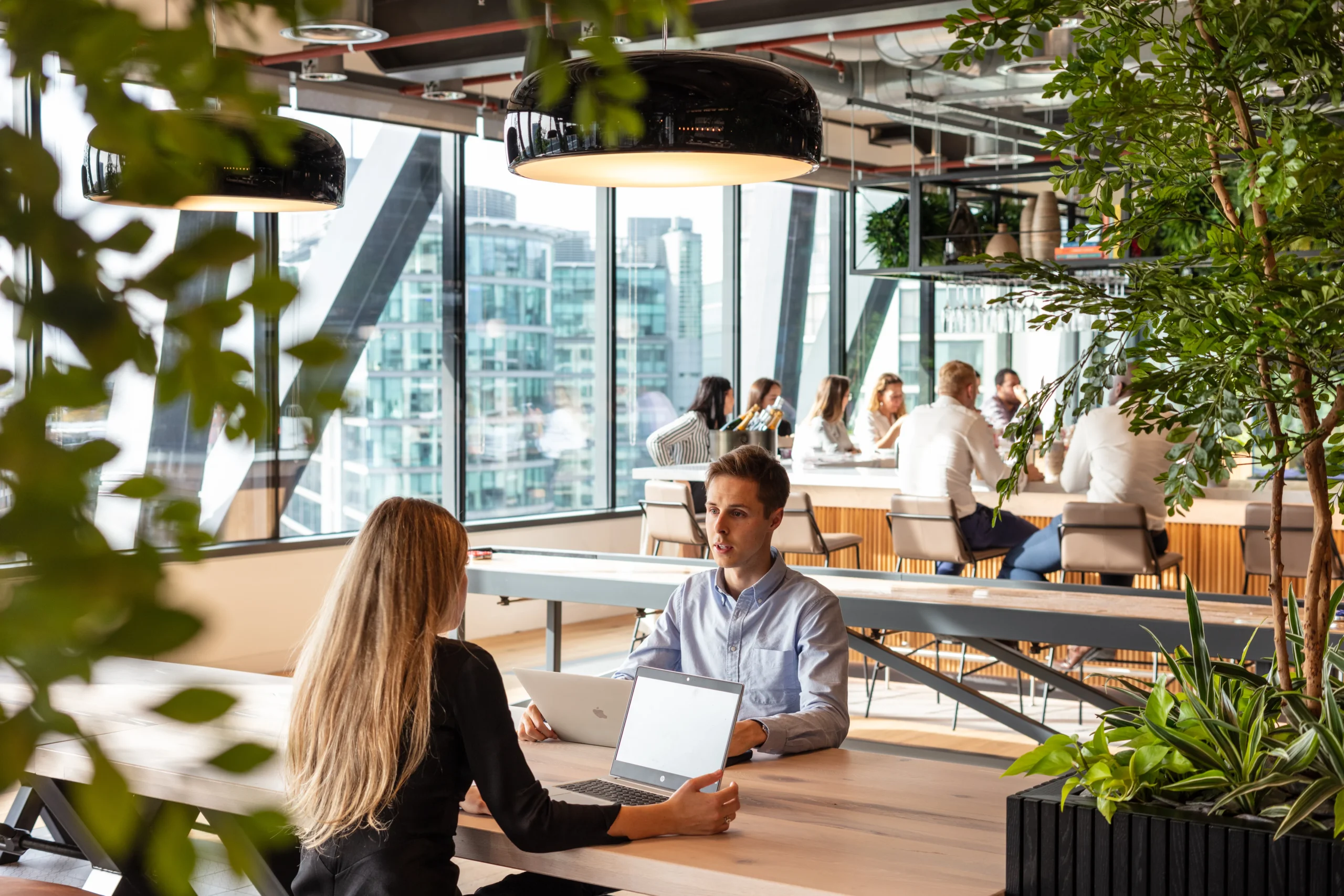 Alpha FX office designed by Modus Workspace, featuring a bright, modern space with large windows and natural light. Two people are seated at a wooden table with laptops, surrounded by greenery and stylish black pendant lighting. In the background, a group of people gather around a communal bar area, fostering collaboration in an open, inviting environment