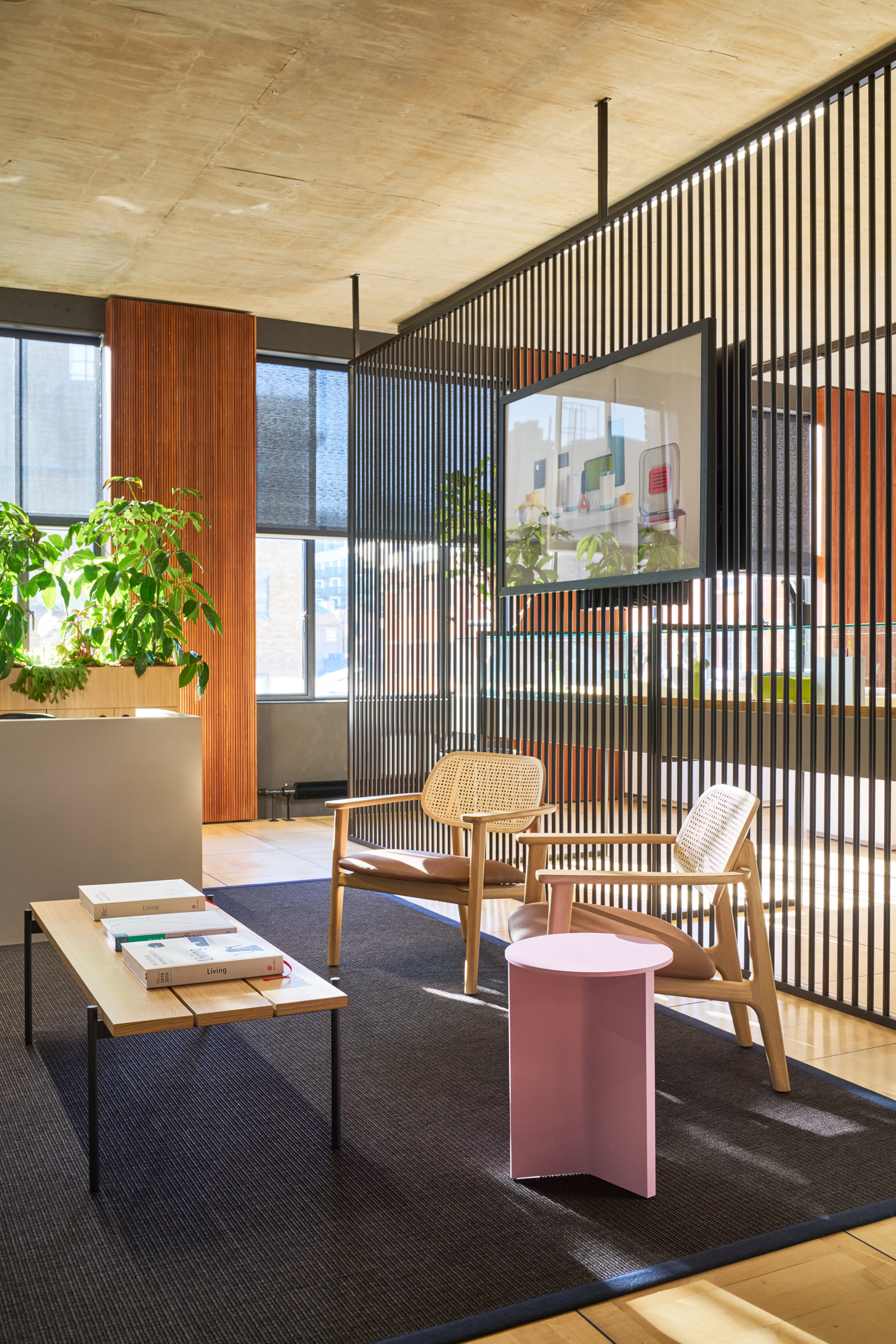 The Joseph Joseph office designed by Modus Workspace features a stylish reception area with wooden accents, modern rattan chairs, and a sleek pink side table. A black slatted divider separates the space, adding privacy while maintaining openness. Greenery and natural light create a welcoming and functional environment for visitors.