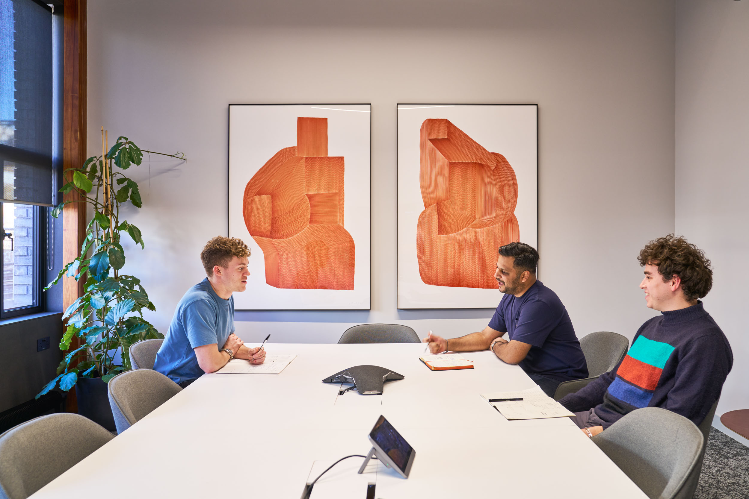 Three individuals in a modern meeting room at Joseph Joseph, designed by Modus, are engaged in a collaborative discussion. The room features contemporary artwork with bold orange designs, a sleek white table, and minimalist decor, creating a bright and productive workspace