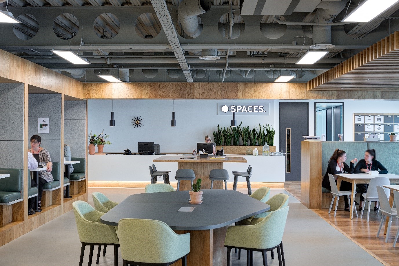 Spaces coworking area designed by Modus Workspace, featuring flexible seating options including booths, collaborative tables, and a reception area. The modern interior is enhanced by natural wood finishes, contemporary lighting, and greenery, offering a welcoming and functional space for work and meetings