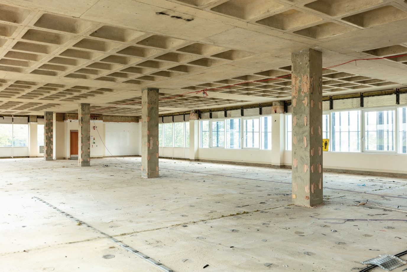 A shell and core office fit-out, showcasing an exposed concrete ceiling grid and unfinished columns in a large, open-plan workspace with large windows providing natural light