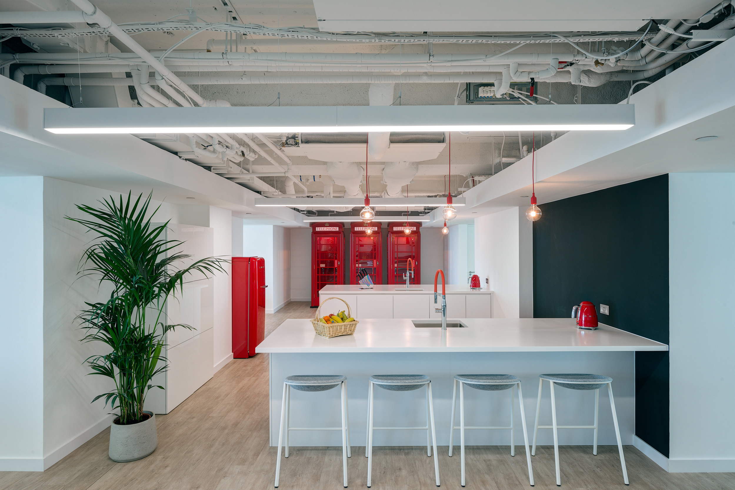 Modern kitchen area in Primalbase's office, designed by Modus Workspace, featuring sleek white surfaces, red accents, and contemporary pendant lighting.