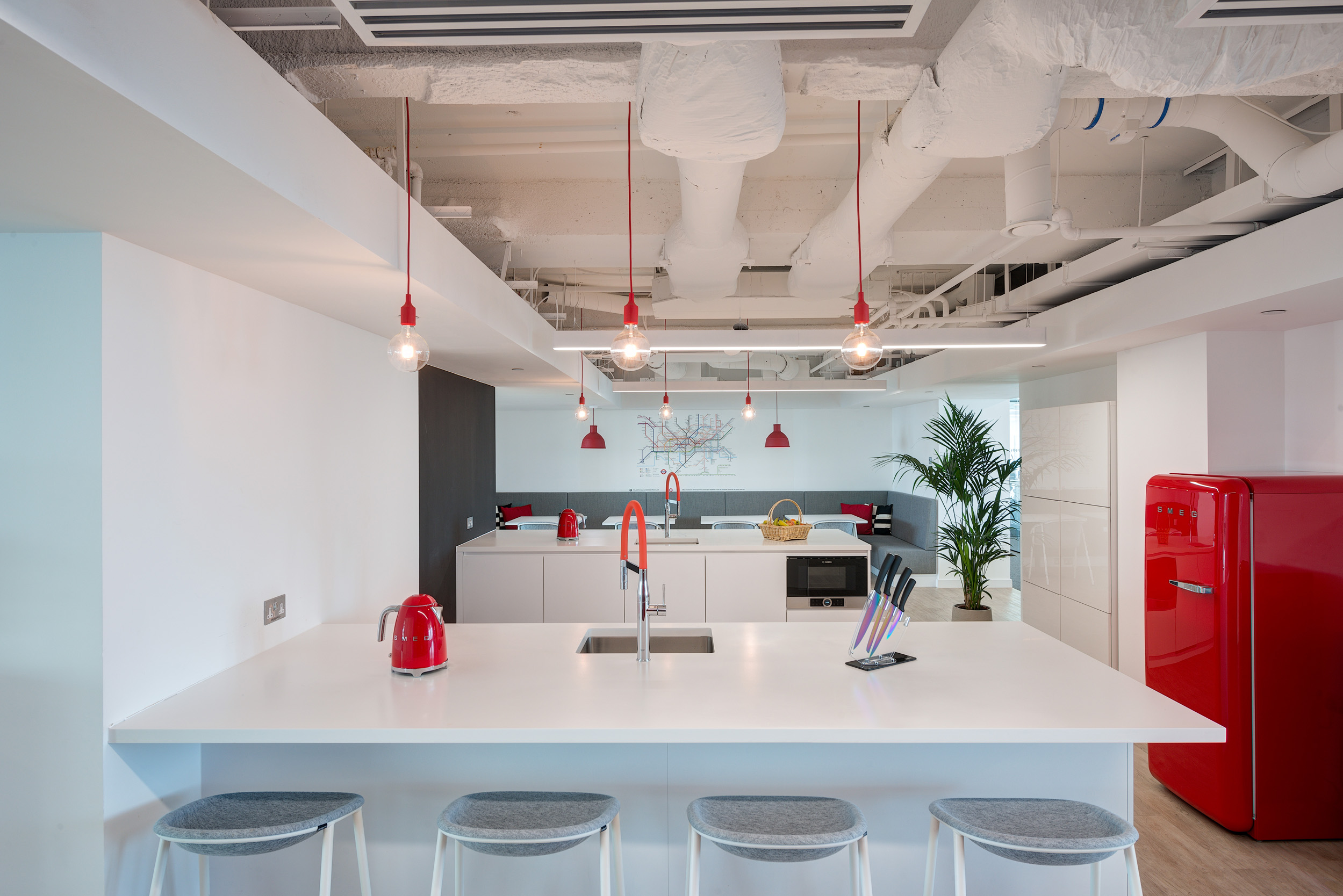Teapoint area in Primalbase's office designed by Modus Workspace, showcasing a minimalist design with white cabinetry, red pendant lights, and a stylish seating area.