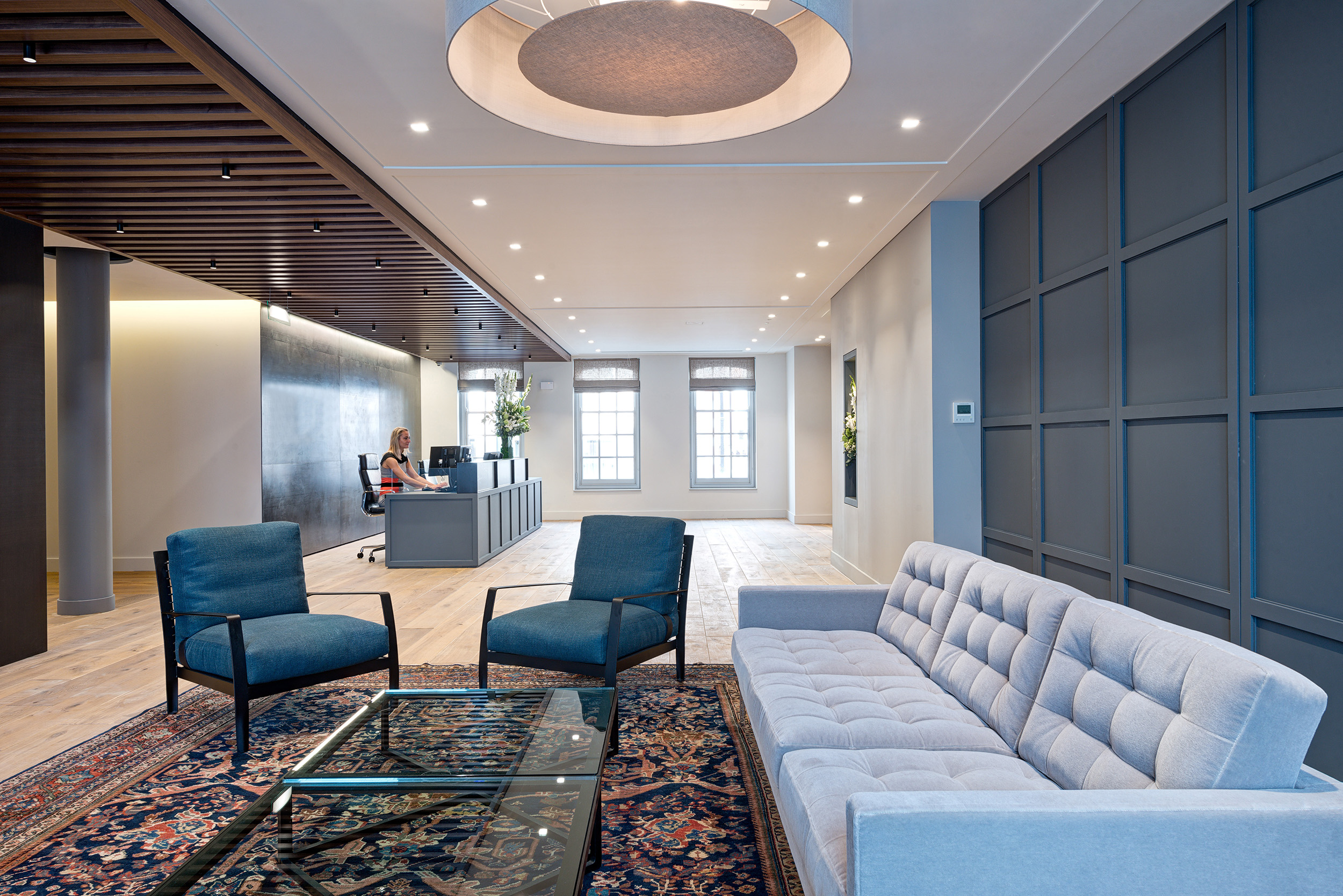 Lounge area in a private equity firm office with blue chairs, grey sofa, and wooden ceiling slats, featuring large windows with natural light.