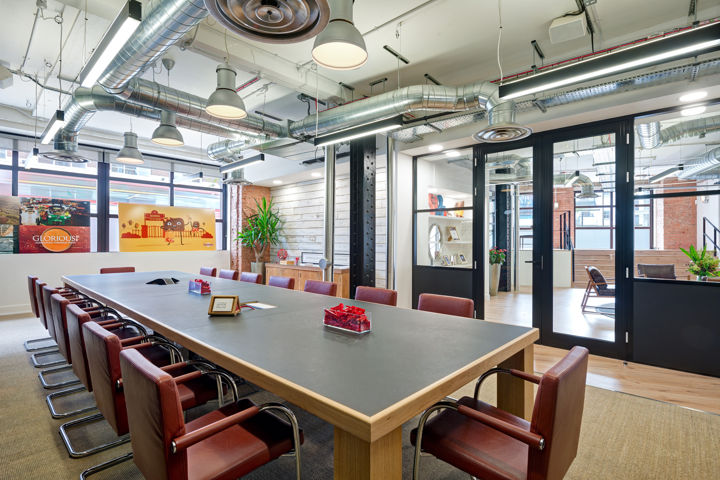 The large meeting room at Red Brick Road office, with industrial elements and exposed ductwork. A large table with red leather chairs and graphic artwork in the windows.
