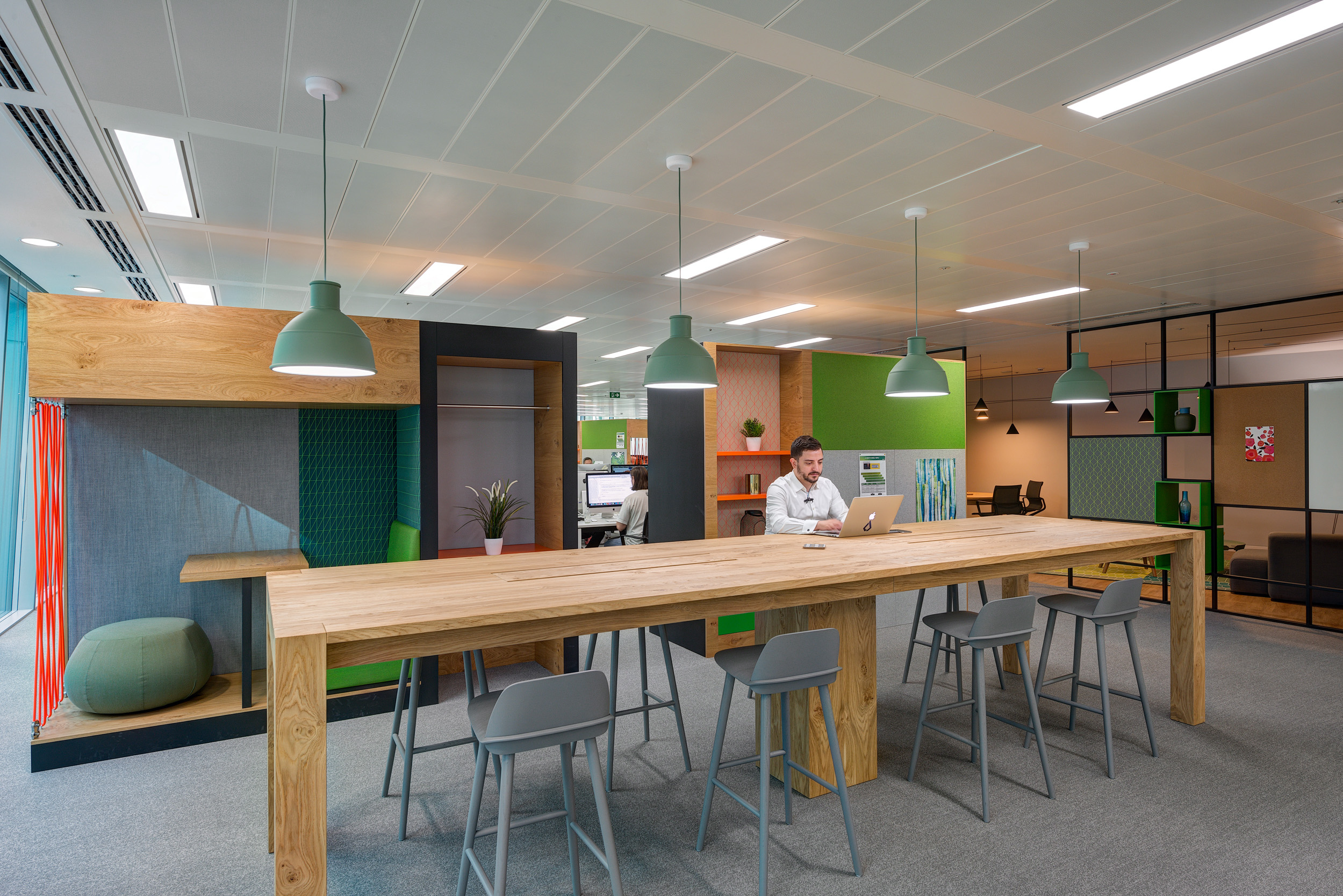 High table workspace in Reply's office designed by Modus Workspace, featuring a large communal wooden table and green pendant lights for informal meetings.