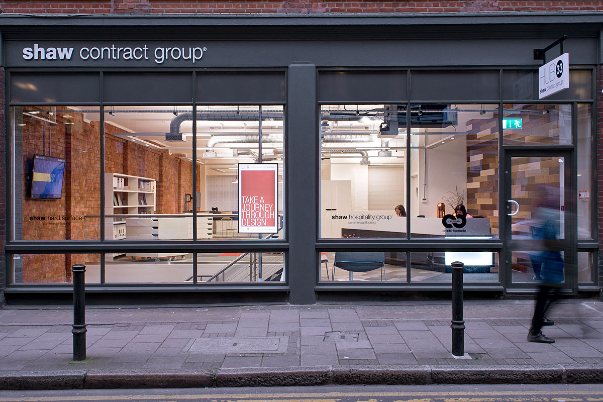 Street-facing exterior of the Shaw Contract Group showroom, showcasing floor-to-ceiling windows and interior design elements with signage promoting their flooring collections.