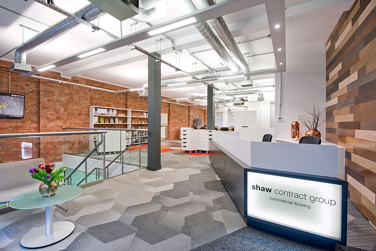 A bright and open reception area at Shaw Contract Group with an industrial-chic design, including a hardwood feature wall and grey geometric carpeting.