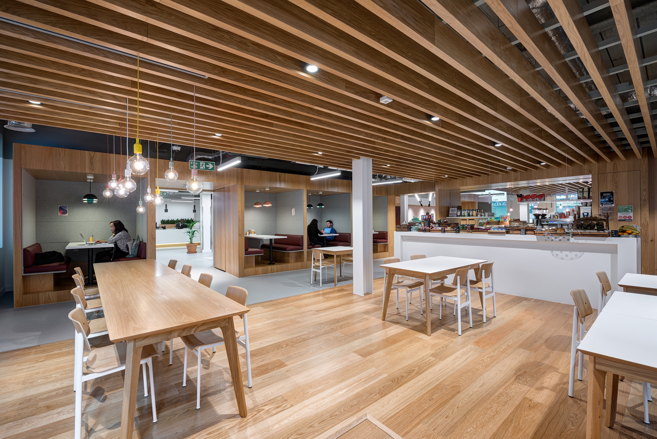 A communal teapoint and working area with long wooden tables, pendant lights, and booth seating at Spaces, 30 Moorgate.