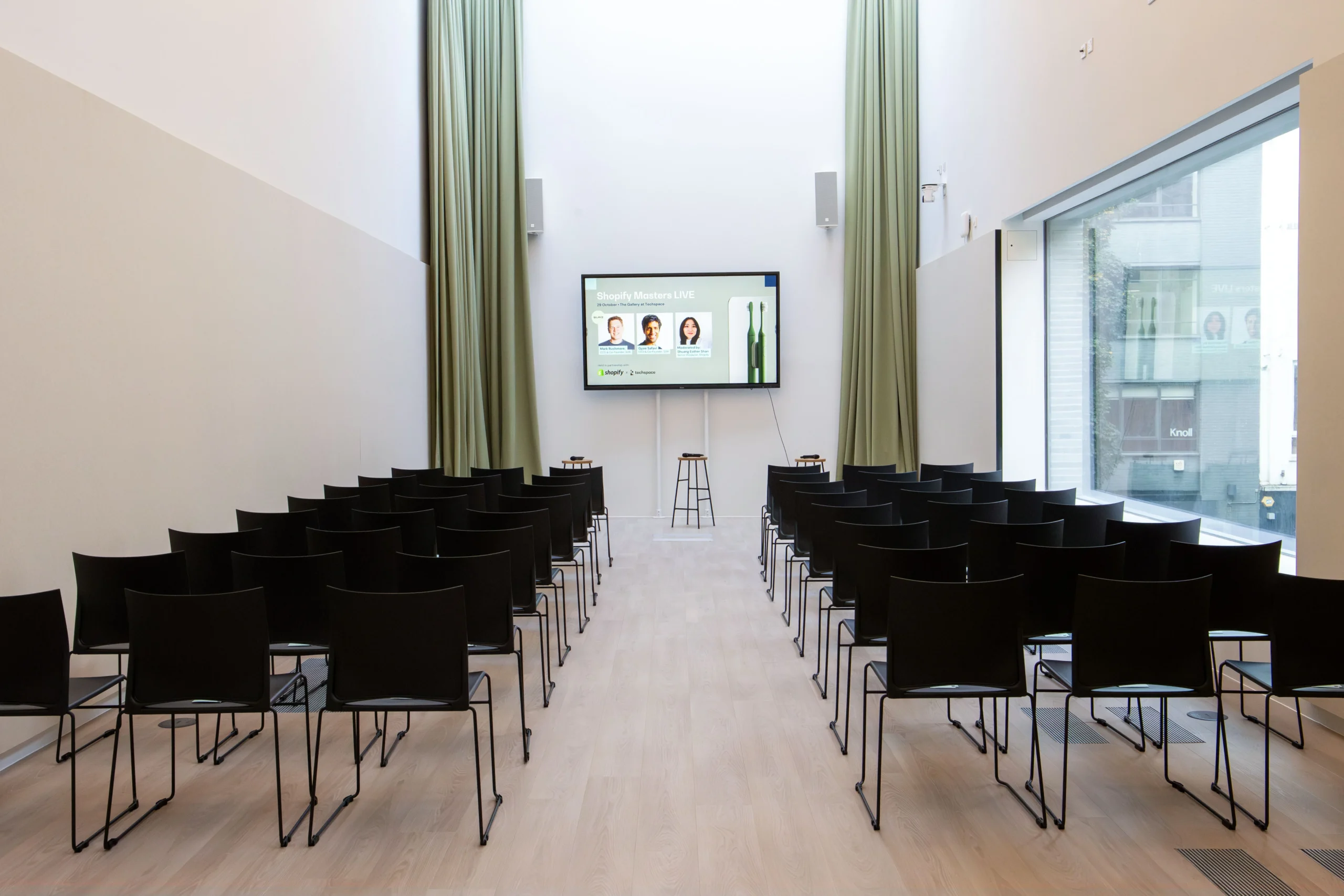 Techspace conference hall, with ceiling to floor sage green curtains, a TV Screen and black seating to view.