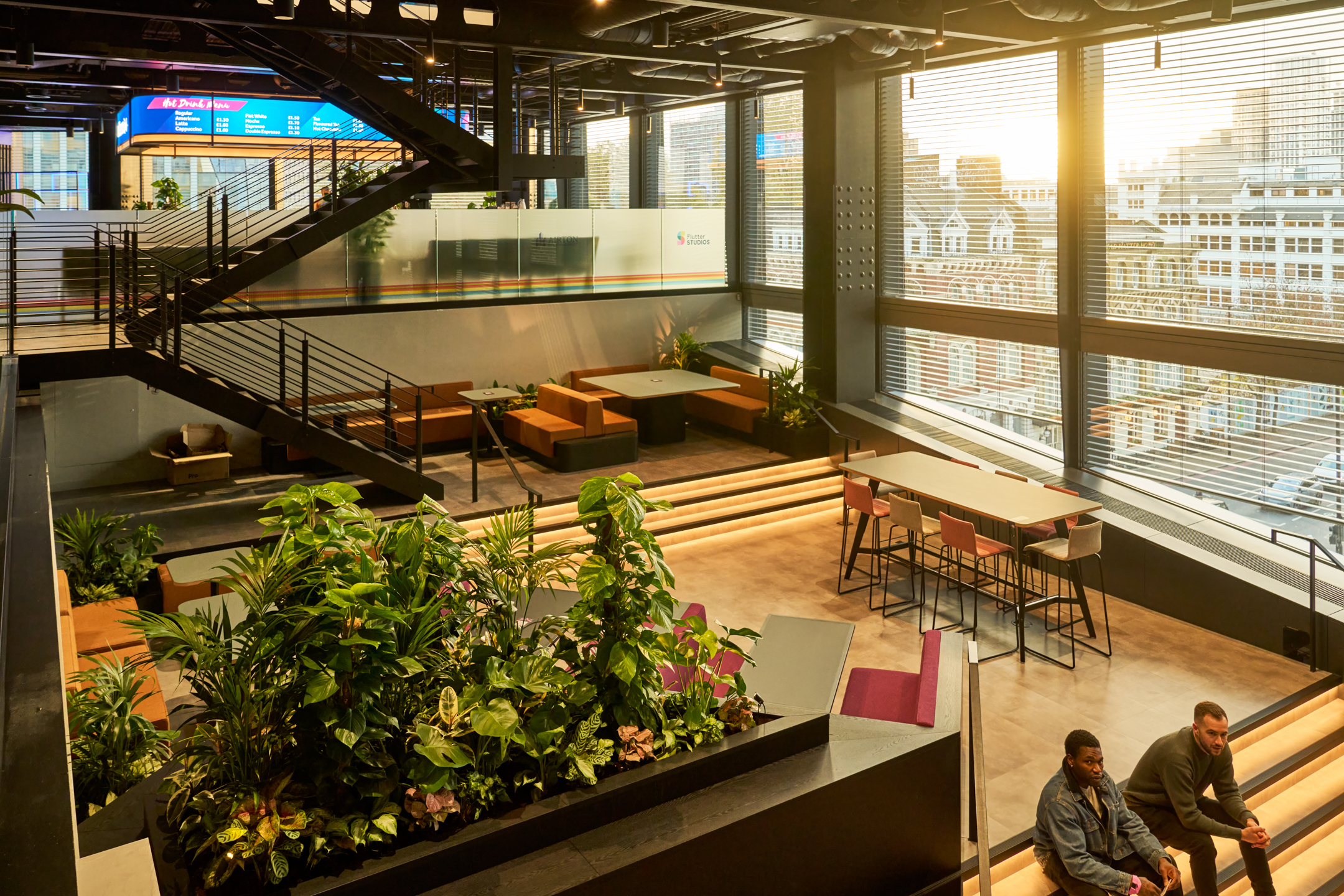 Sunlit atrium space inside an Entertainment Company office, featuring tiered seating, lush planting, and collaborative booths overlooking the city.