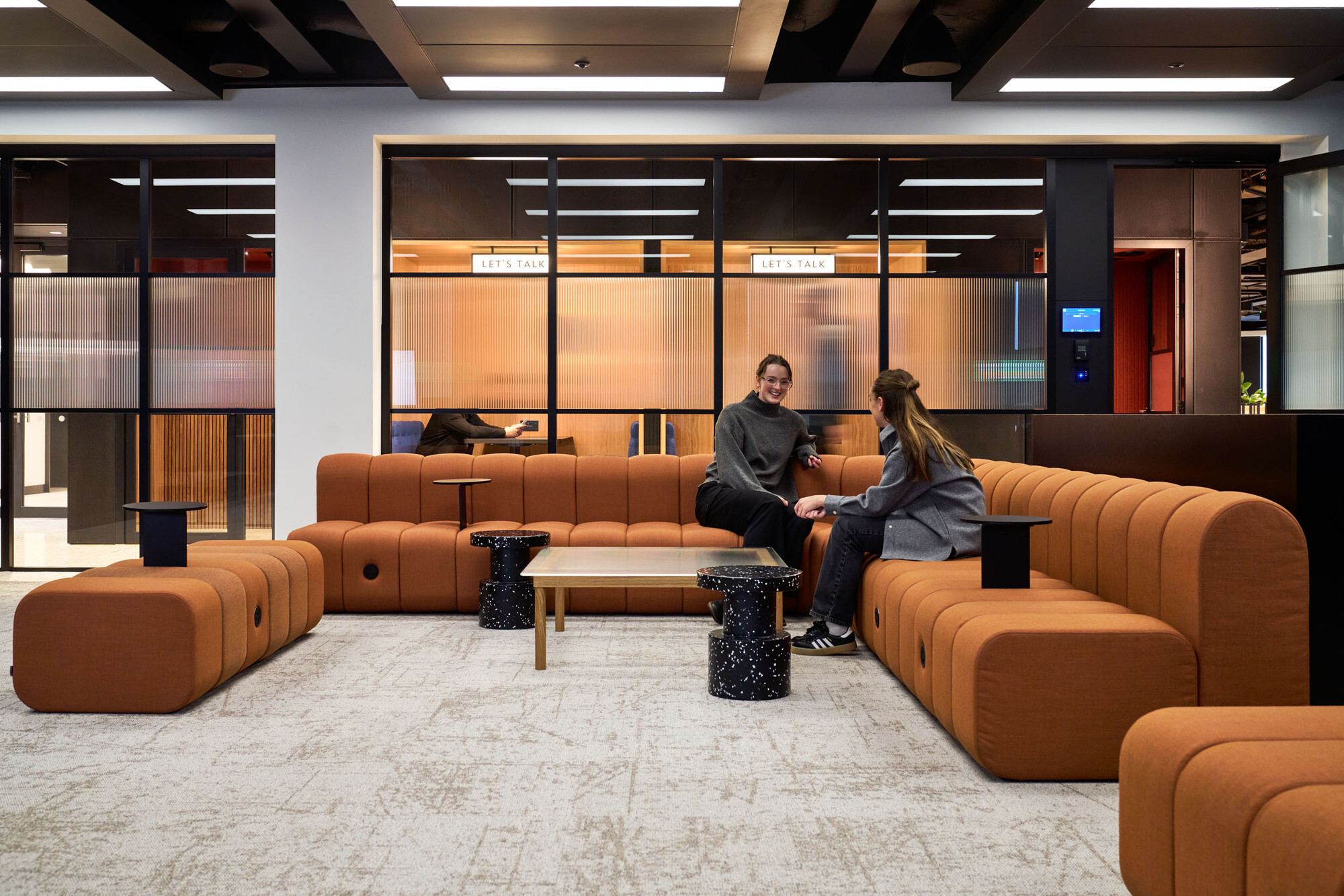 International Technology and Data Firm employees happily having a catch up on the large orange sofa, in front of two glass meeting rooms.
