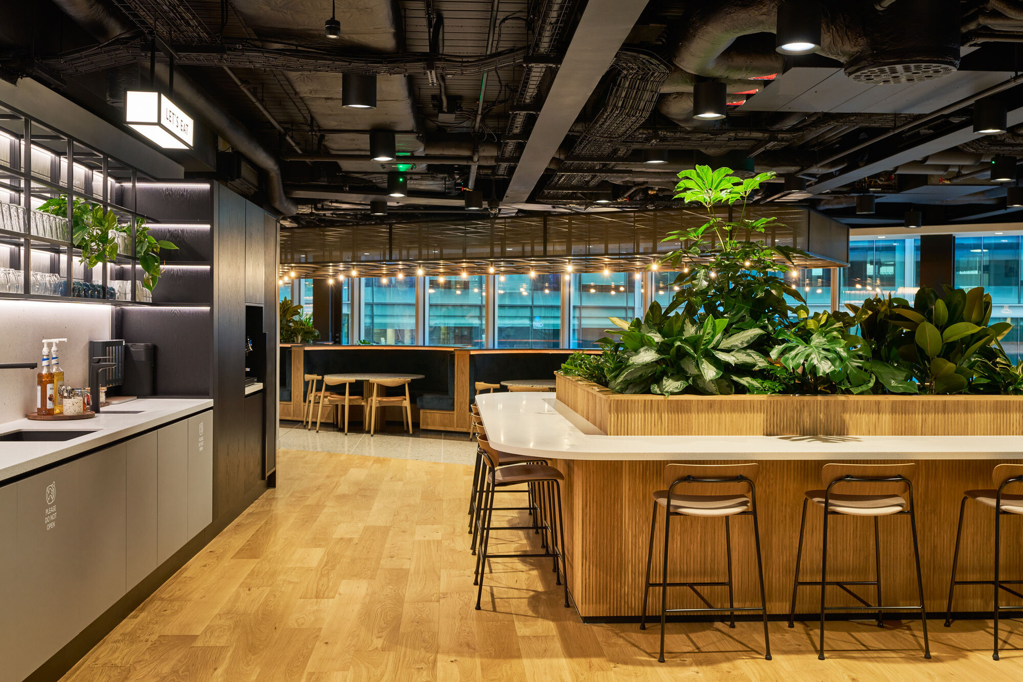 International Technology and Data Firm teapoint, designed by Modus, featuring a large central point of plotted plants, an exposed black painted ceiling, kitchen utensils and an island with high stools.