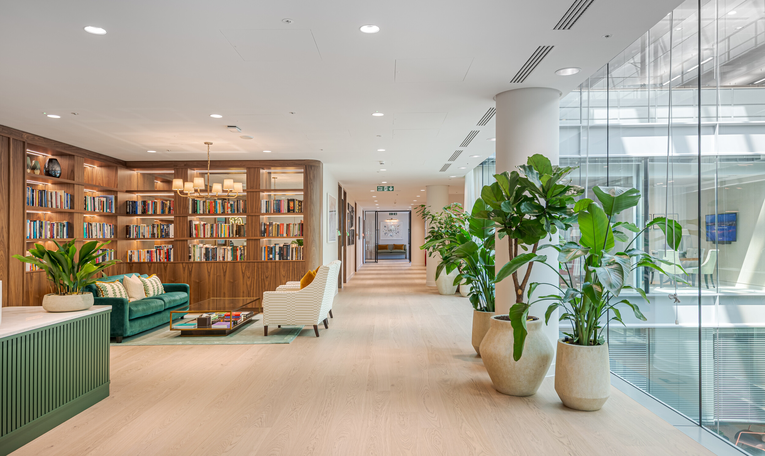 Library-style breakout space in an Investment Management Firm office fit out by Modus. Includes walnut bookshelves, a velvet green sofa, and biophilic elements. Floor-to-ceiling glazing brings natural light into the corridor.