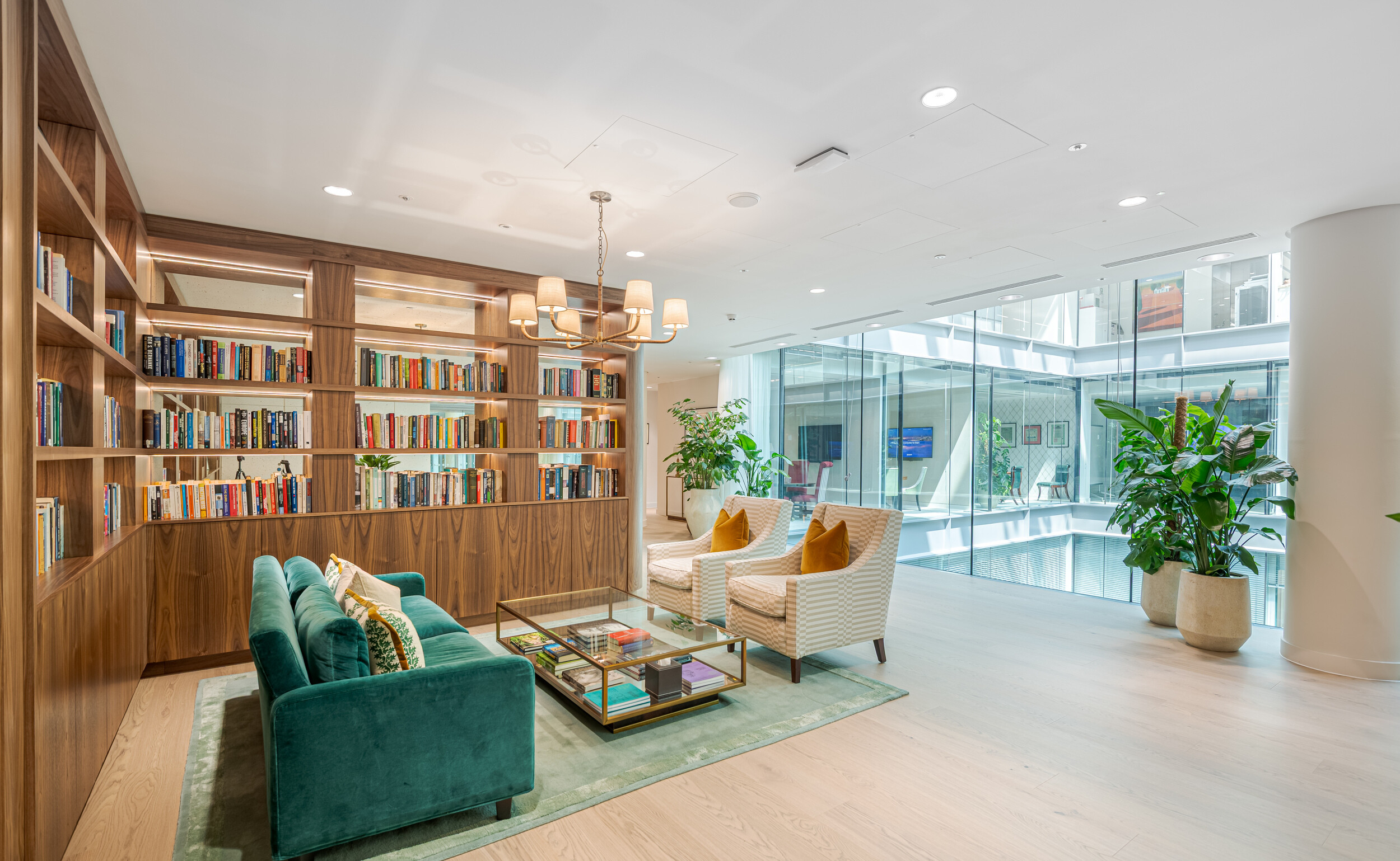 Library corner in an Investment Management Firm office fit out by Modus, featuring open walnut shelving and curated titles. A velvet green sofa, striped armchairs, and layered textures create a boutique hotel feel. Floor-to-ceiling glazing overlooks an internal atrium, drawing in daylight.
