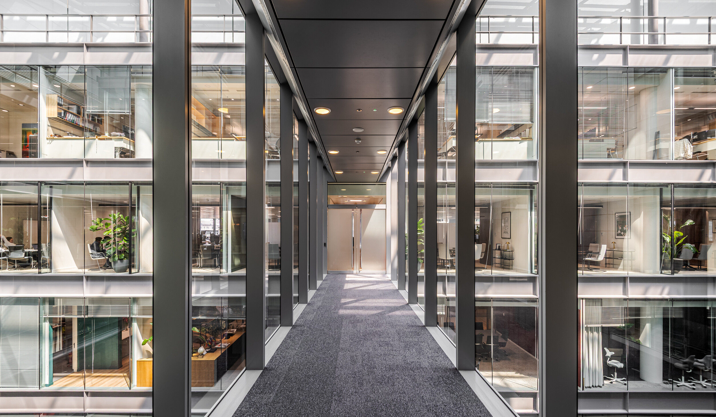 Glazed bridge walkway at the centre of an Investment Management Firm office, connecting workspace zones across an atrium. Surrounded by multiple levels of glass-fronted meeting rooms and desking. A striking architectural feature within the Modus-designed fit out.