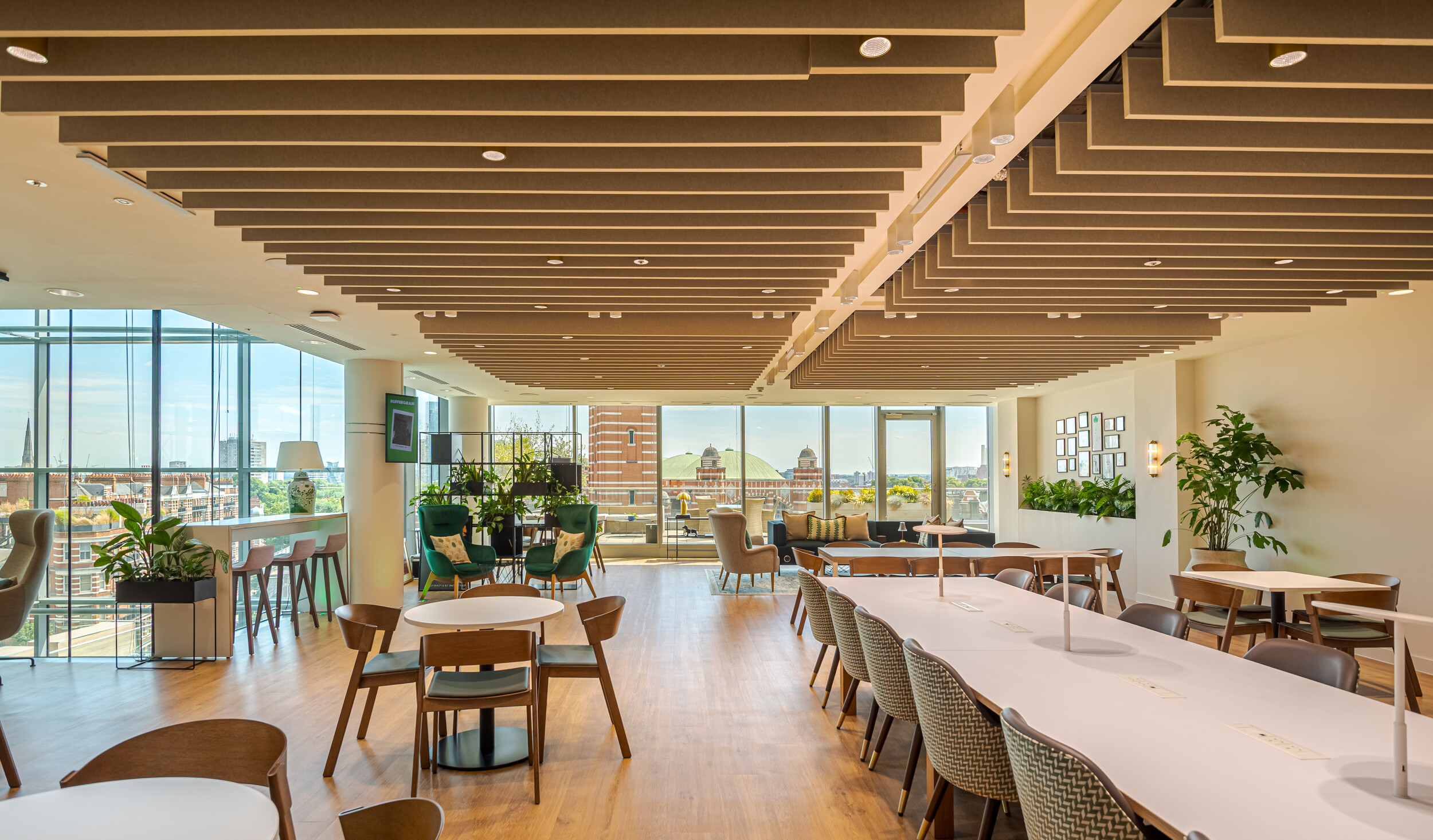 Communal lounge and breakout space within an Investment Management Firm office, designed by Modus. Acoustic ceiling baffles, warm timber flooring, and panoramic city views create a welcoming atmosphere. A mix of soft seating, dining tables and focus benches supports agile working.