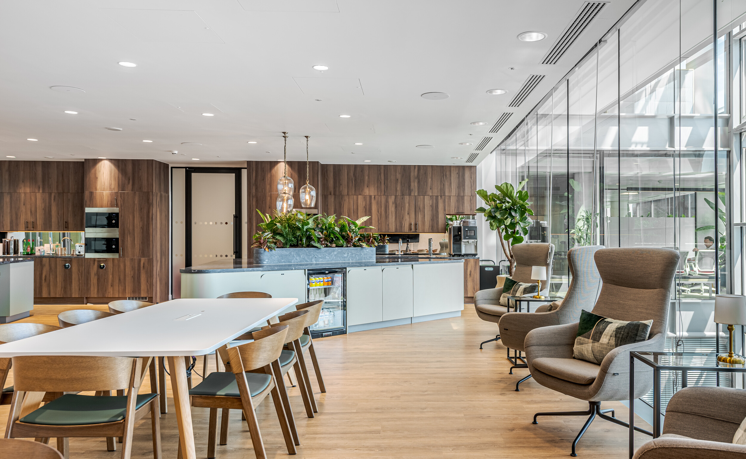 Teapoint and social hub in an Investment Management Firm office fit out by Modus, featuring walnut cabinetry and a central island with built-in planting. Lounge chairs line the glazed wall, offering a quiet retreat. A balance of comfort and functionality in a refined shared space.