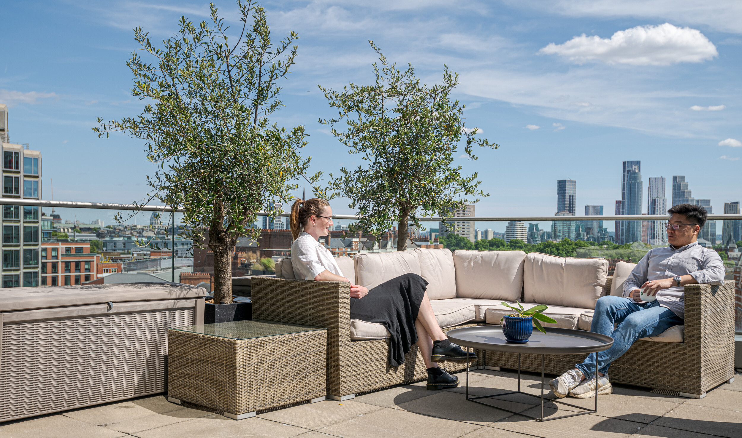 Rooftop terrace at an Investment Management Firm with rattan lounge furniture and skyline views. Two employees relax in the sunshine beneath olive trees in planters. A calm, open-air setting designed for informal catch-ups and fresh air.