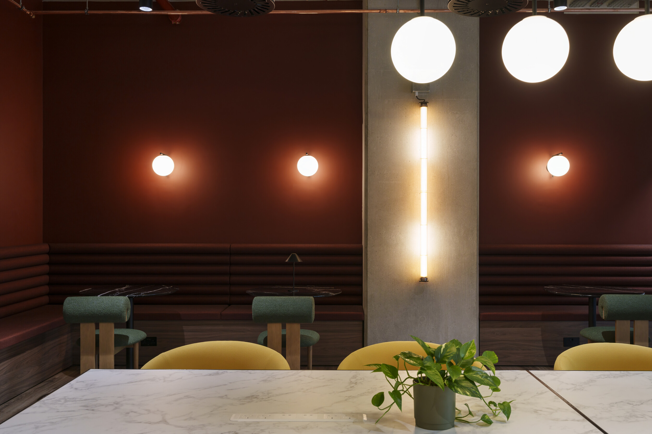 Warm-toned seating area at Newflex One Station Hill with burgundy banquettes, green chairs, and globe lighting, viewed from a marble communal table with plants.