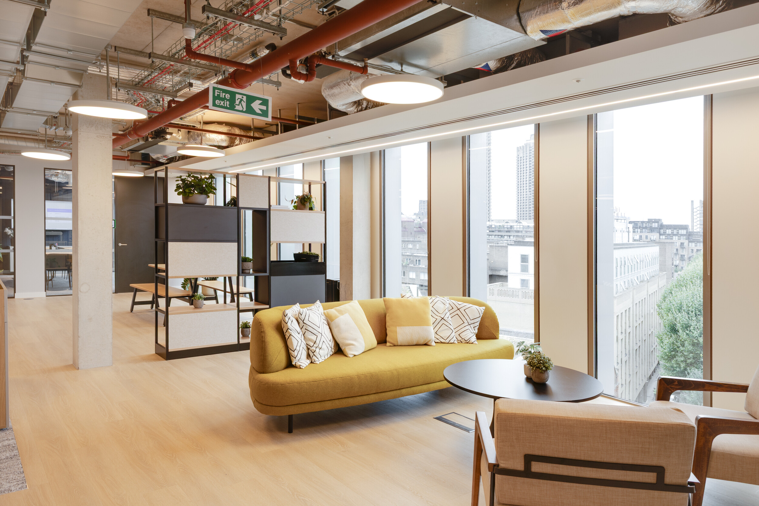 Capital on Tap breakout space with mustard sofa, modern lounge seating, shelving with plants, and floor-to-ceiling windows overlooking the city.