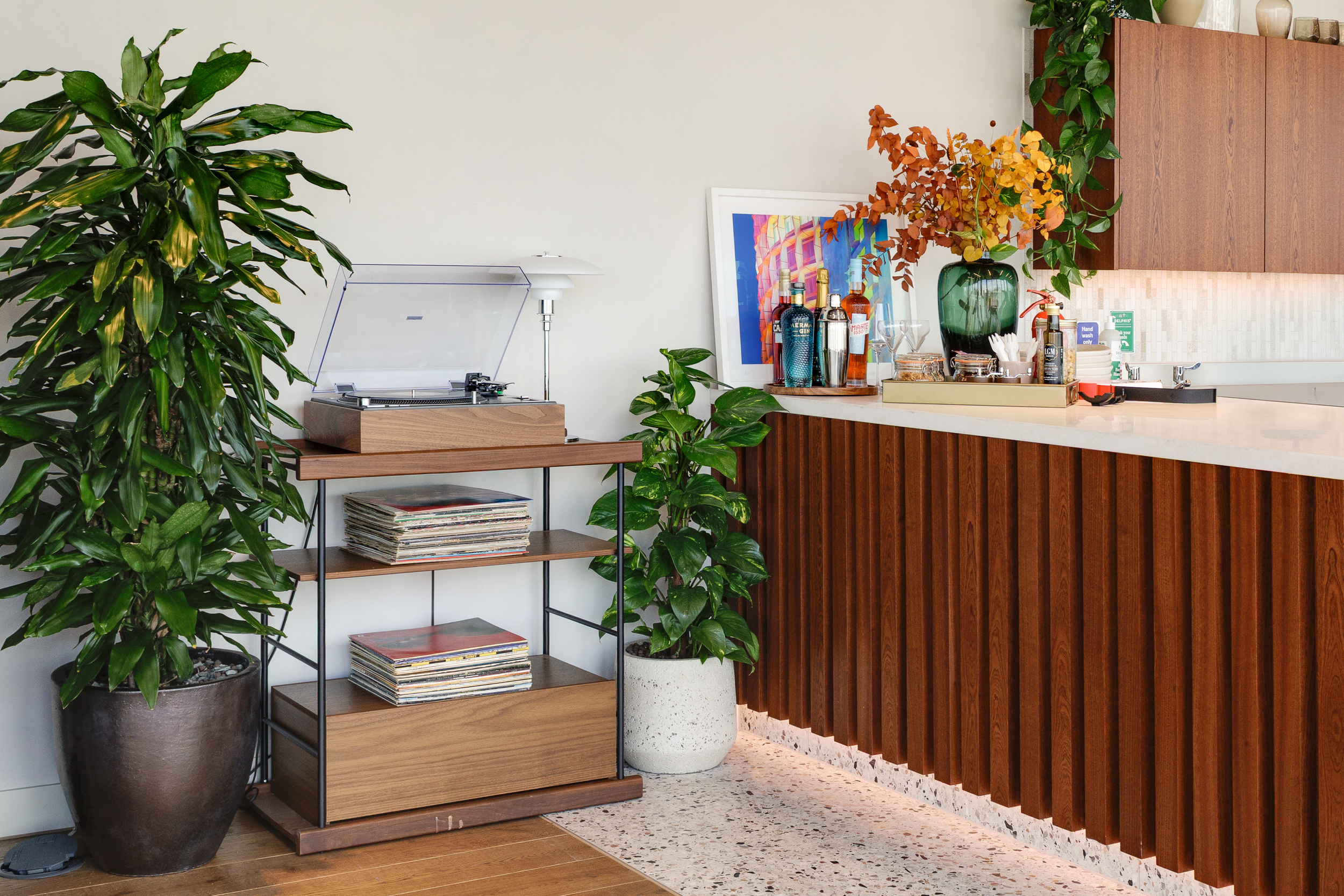 Corner detail of the Space House Clubhouse showing a walnut-fluted bar, terrazzo flooring, and a vinyl station surrounded by lush greenery, part of Modus’ interior design.