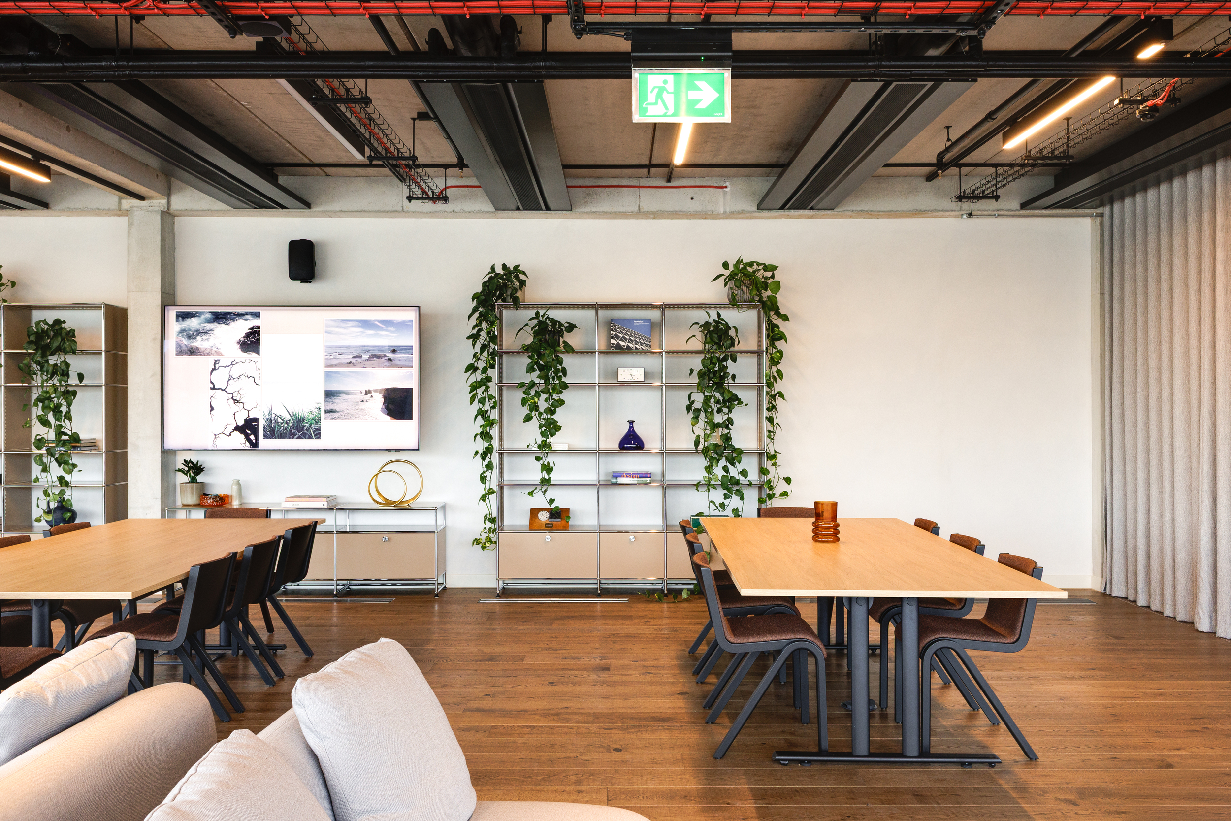 Collaborative area inside the Space House Clubhouse, designed by Modus, with long timber tables, shelving decorated with greenery, and wall-mounted digital displays.