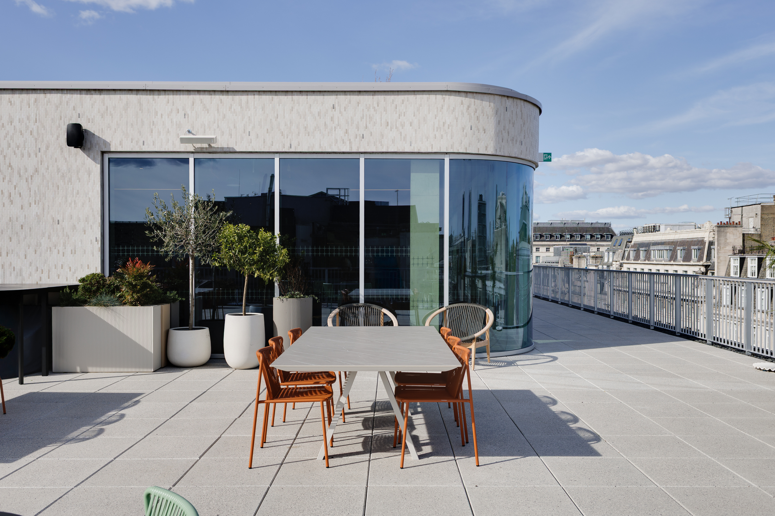 The Space House Clubhouse terrace with a mix of orange and neutral furniture, large planters, and curved glass façades reflecting London’s skyline, completed by Modus.